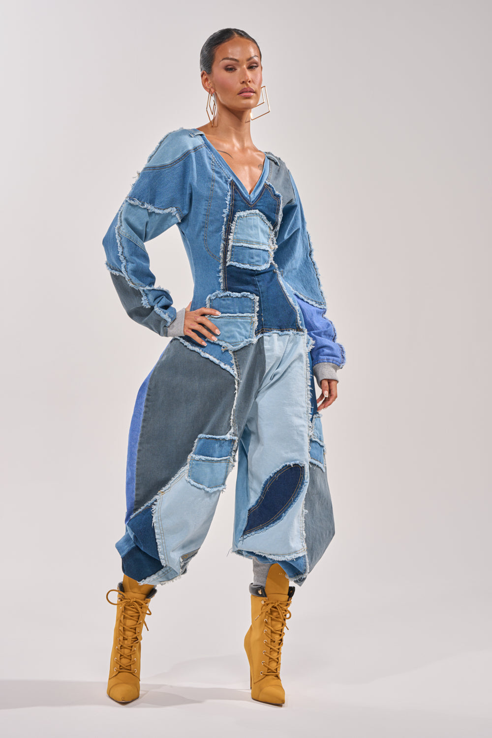 A person poses confidently in the COMFY COZY JUMPSUIT IN DENIM PATCHWORK, featuring various blue hues and raw edges, paired with large hoop earrings and mustard yellow lace-up boots against a plain light background. 0