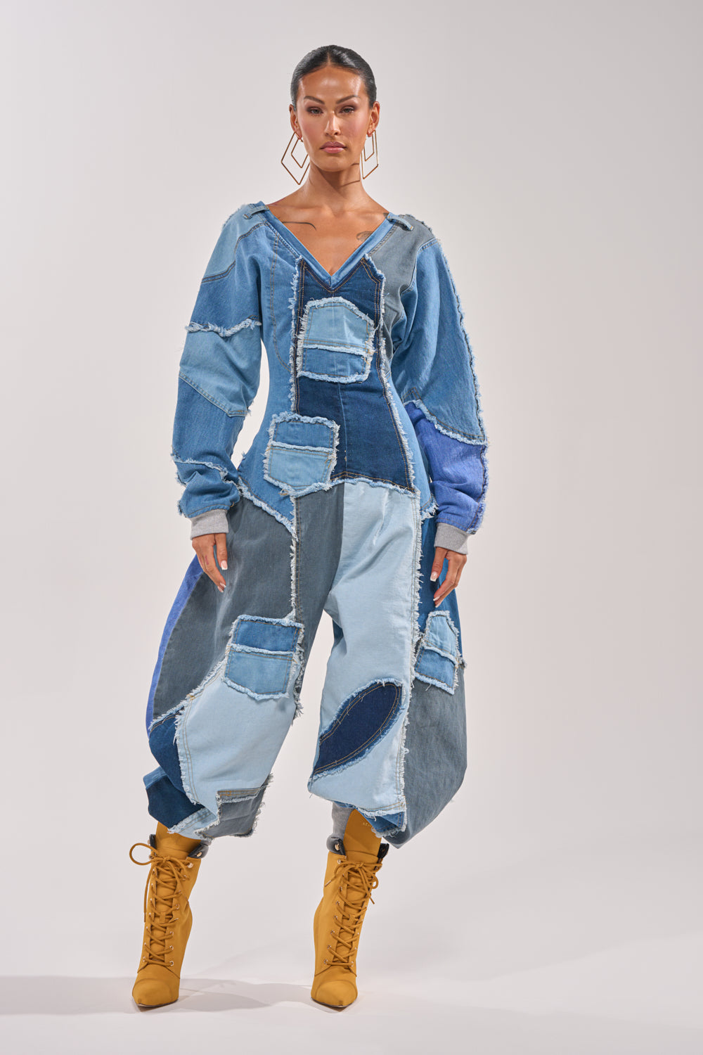 A woman exudes confidence in the COMFY COZY JUMPSUIT IN DENIM PATCHWORK, featuring oversized patchwork denim in blue tones with raw edges, styled with tan lace-up heeled boots and geometric earrings for a bold streetwear look. 1
