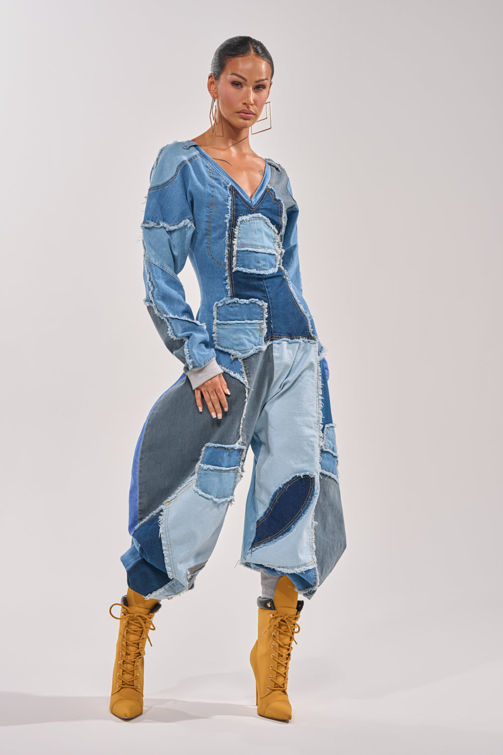 A woman models the COMFY COZY JUMPSUIT IN DENIM PATCHWORK, featuring mixed blue tones and frayed edges. Paired with tan lace-up boots and hoop earrings, she stands confidently against a light background radiating streetwear style. 2