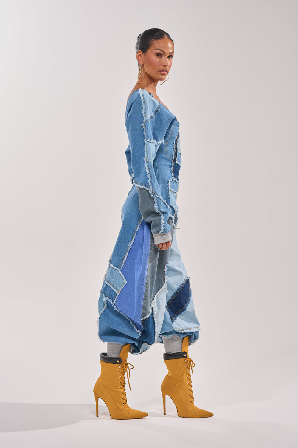 A woman in profile wears the COMFY COZY JUMPSUIT IN DENIM PATCHWORK, featuring light, medium, and dark blue sections with frayed edges. She styles this trendy piece with tan lace-up high-heeled ankle boots against a white background. 3