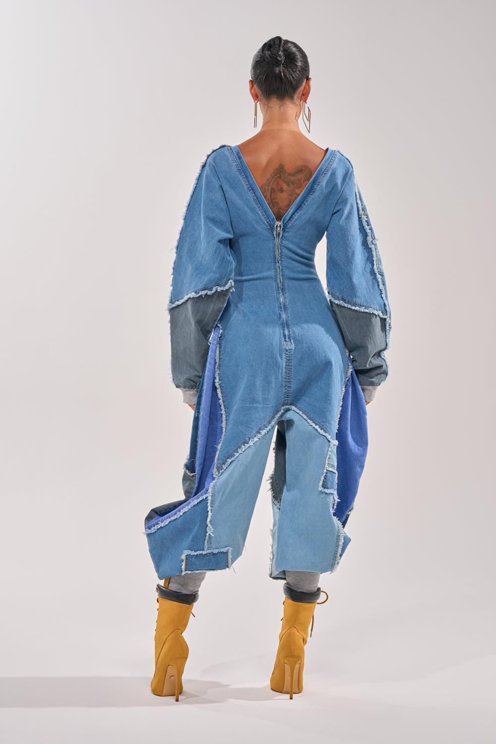 A person with braided hair and a back tattoo stands facing away, wearing the COMFY COZY JUMPSUIT IN DENIM PATCHWORK with frayed edges and dramatic sleeves, paired with yellow heeled boots against a plain background. 5