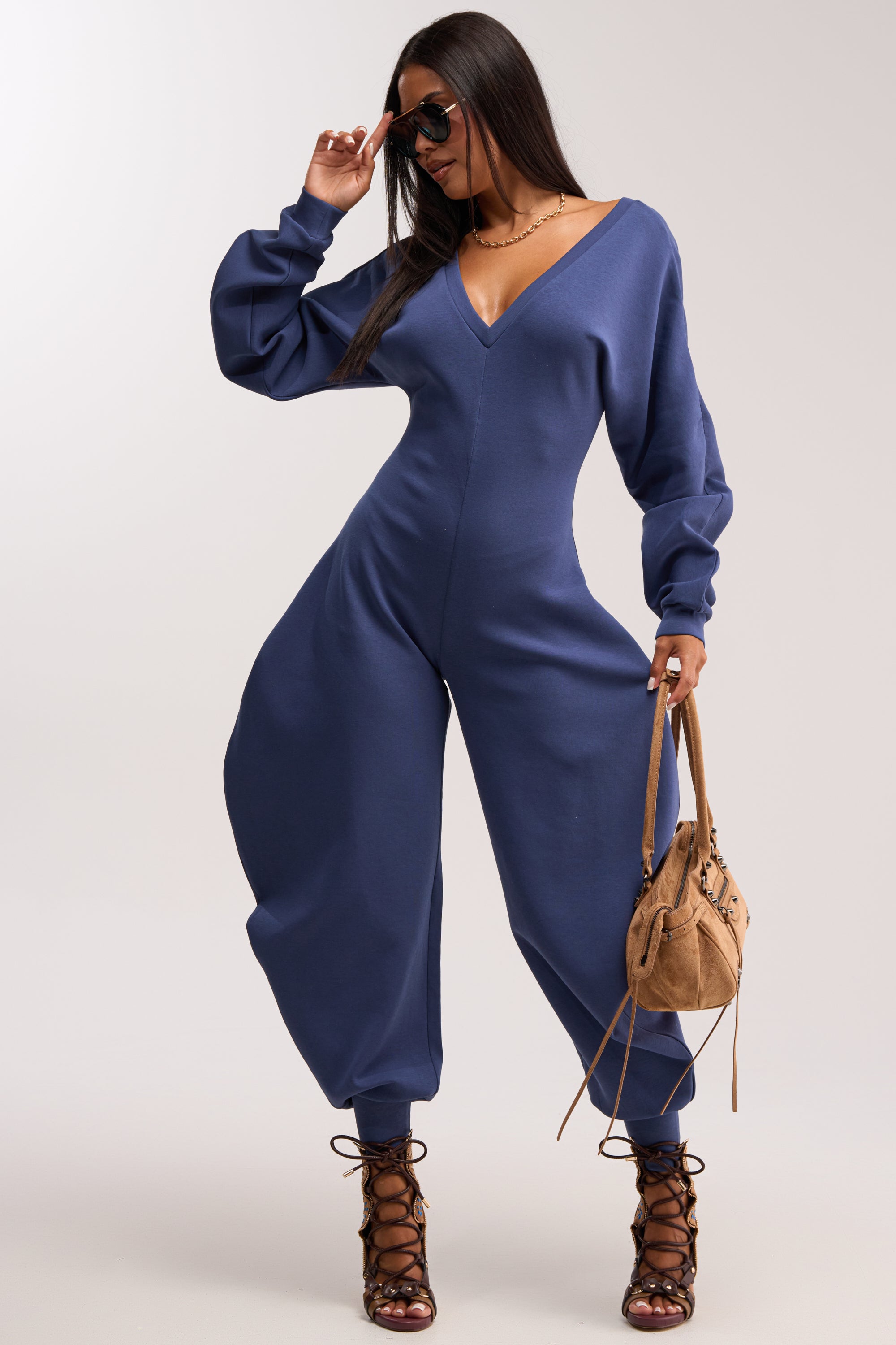 A woman poses confidently in the COMFY COZY SWEATSHIRT JUMPSUIT IN NAVY, featuring wide legs and a fitted waist, styled with a tan handbag, lace-up heels, and sunglasses—a fresh twist on contemporary streetwear.