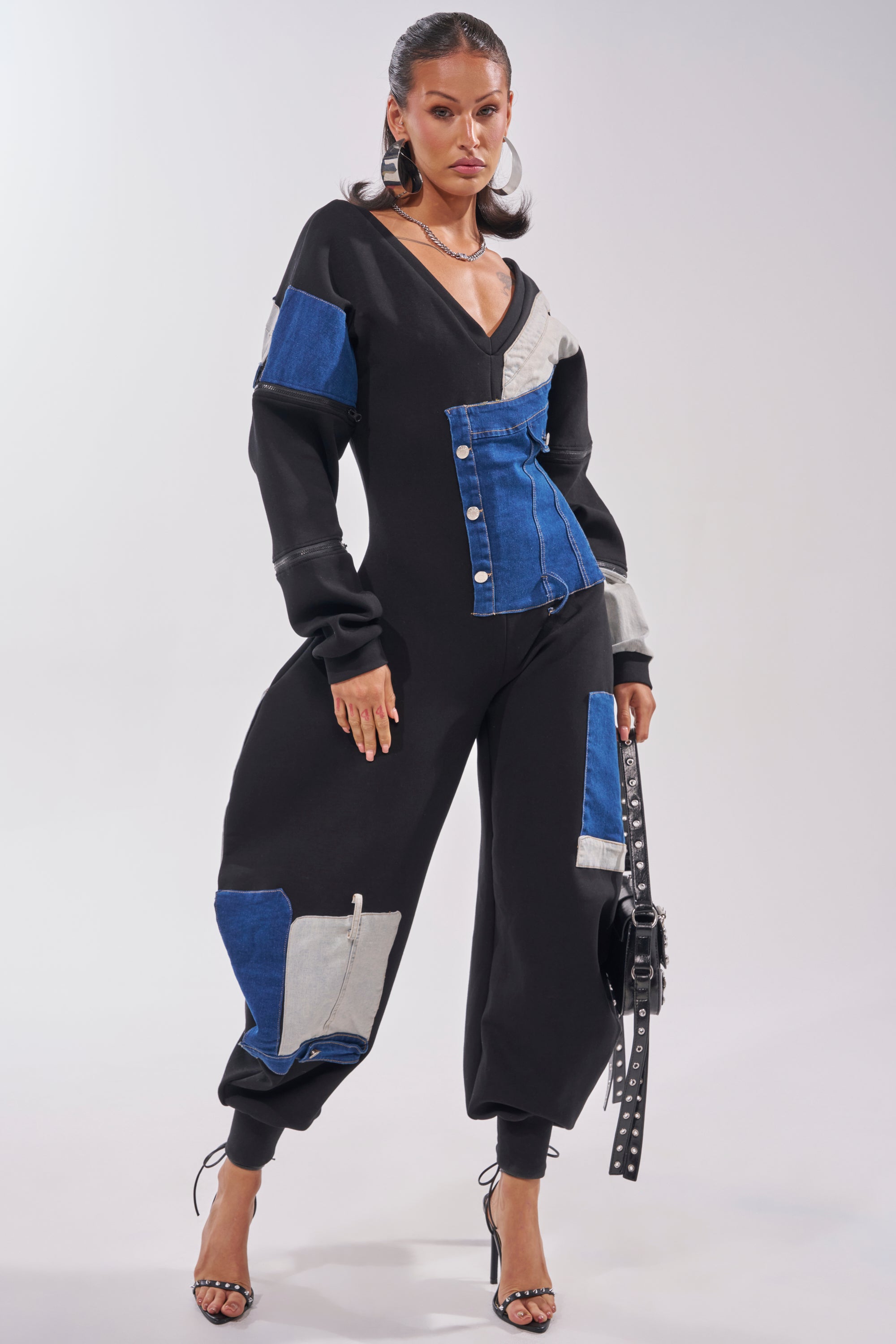 A woman poses confidently in strappy high heels, holding a black belt while wearing the COMFY COZY SWEATSHIRT JUMPSUIT IN PATCHWORK DENIM IN BLACK. 0