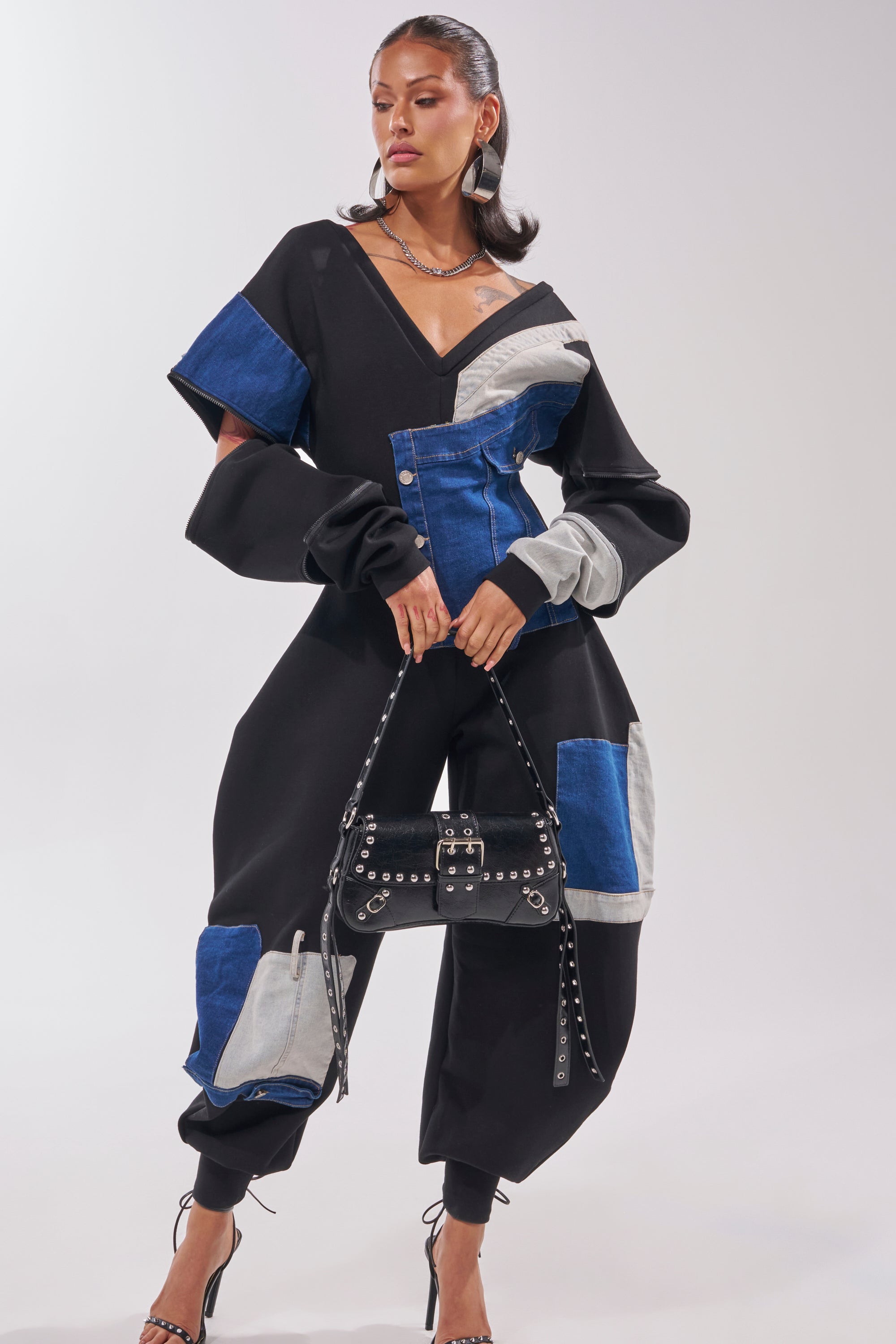 A woman poses in the COMFY COZY Sweatshirt Jumpsuit in Patchwork Denim in Black, featuring black, blue, and gray panels, accessorized with a studded handbag, strappy heels, and layered necklaces against a plain light background. 1