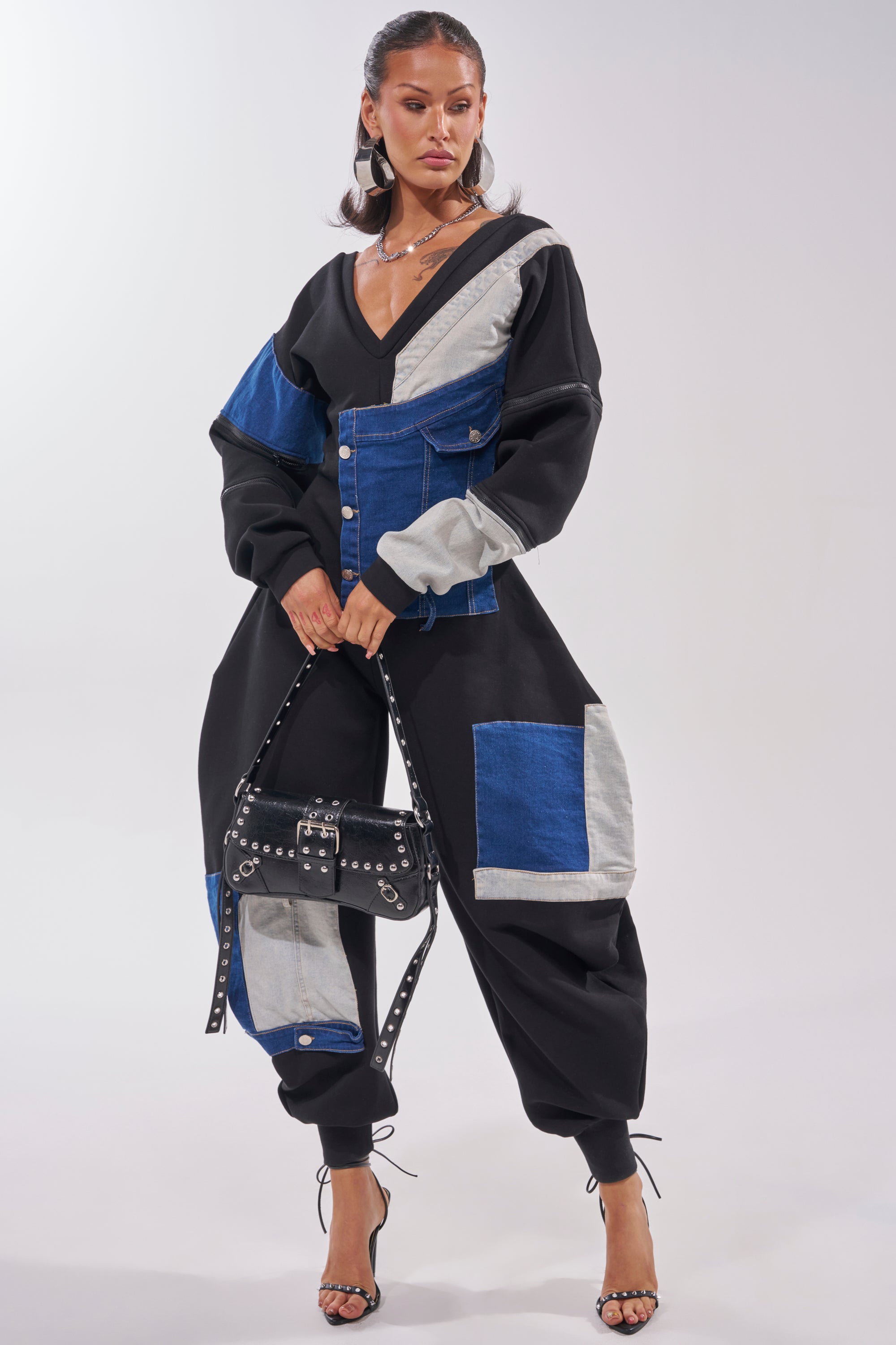 A woman poses in the COMFY COZY SWEATSHIRT JUMPSUIT IN PATCHWORK DENIM IN BLACK, featuring blue, black, and gray panels, paired with a black studded handbag and high-heeled sandals against a plain background. 2