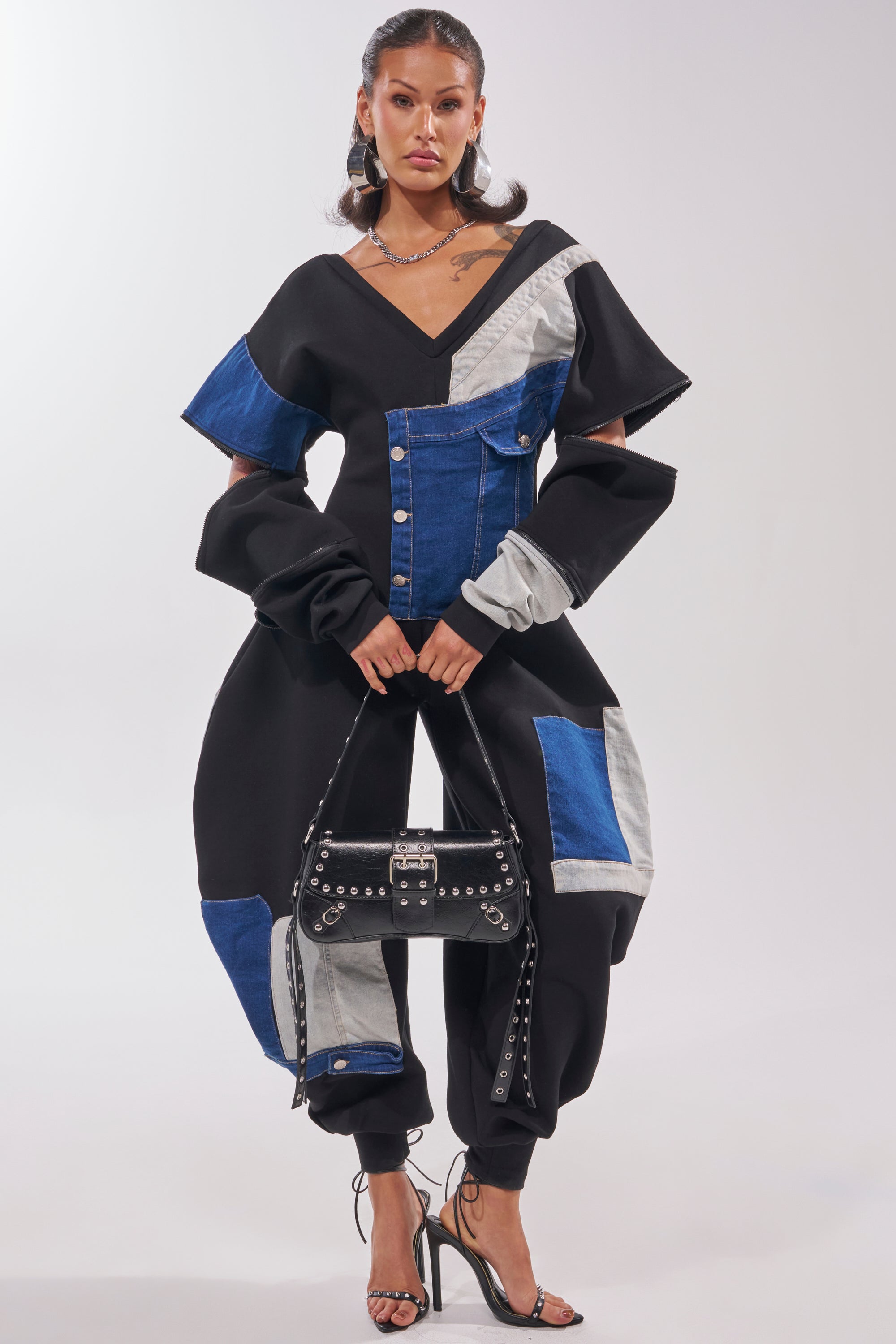 Woman wearing the COMFY COZY SWEATSHIRT JUMPSUIT IN PATCHWORK DENIM IN BLACK with blue and gray denim patches, structured sleeves and pants, holding a studded black handbag and strappy high-heeled sandals against a plain background. 3