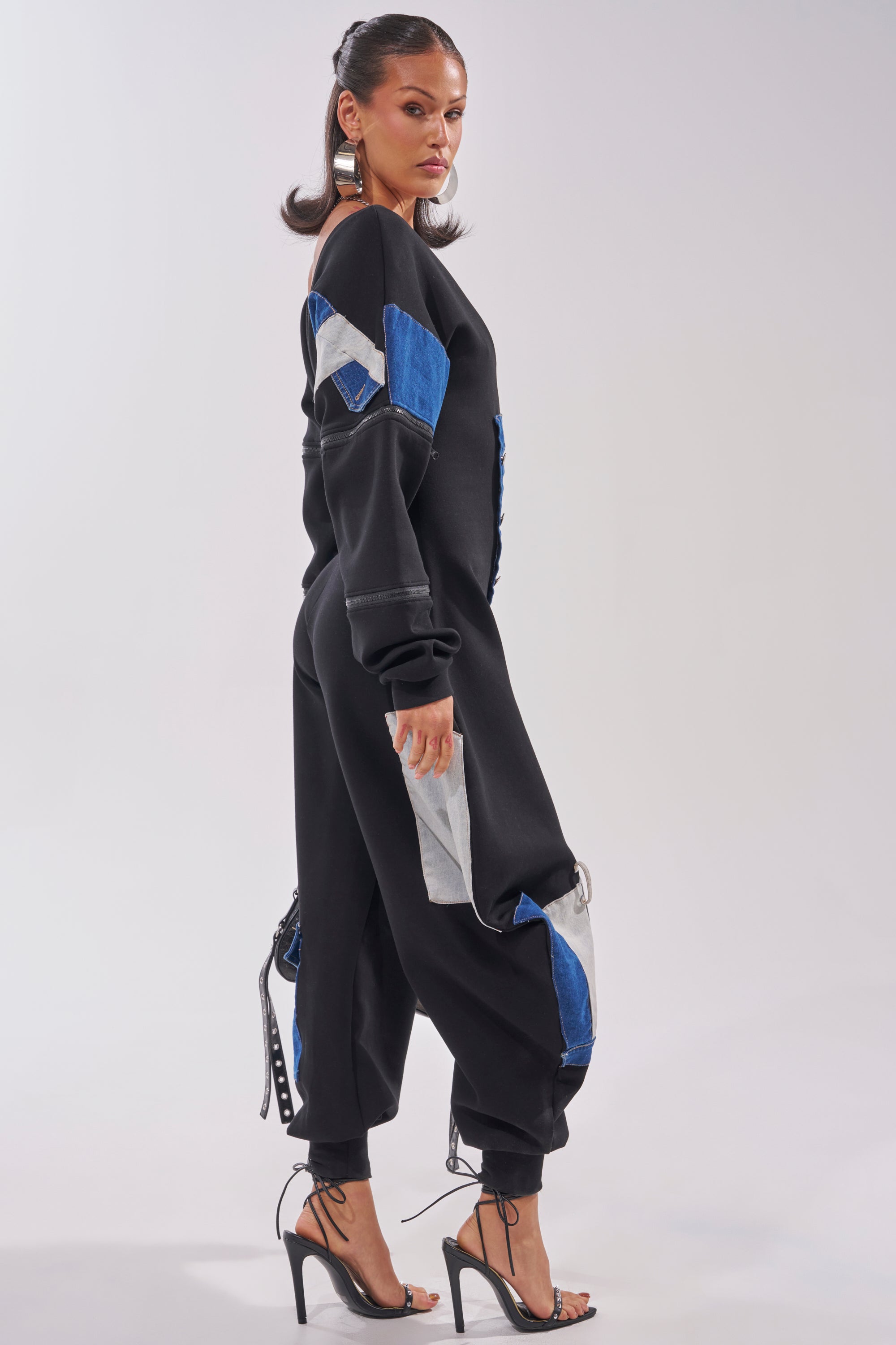 A woman stands sideways wearing the COMFY COZY SWEATSHIRT JUMPSUIT IN PATCHWORK DENIM IN BLACK with jogger cuffs and blue-gray details, styled with high-heeled sandals and a sleek ponytail for streetwear vibes. 4