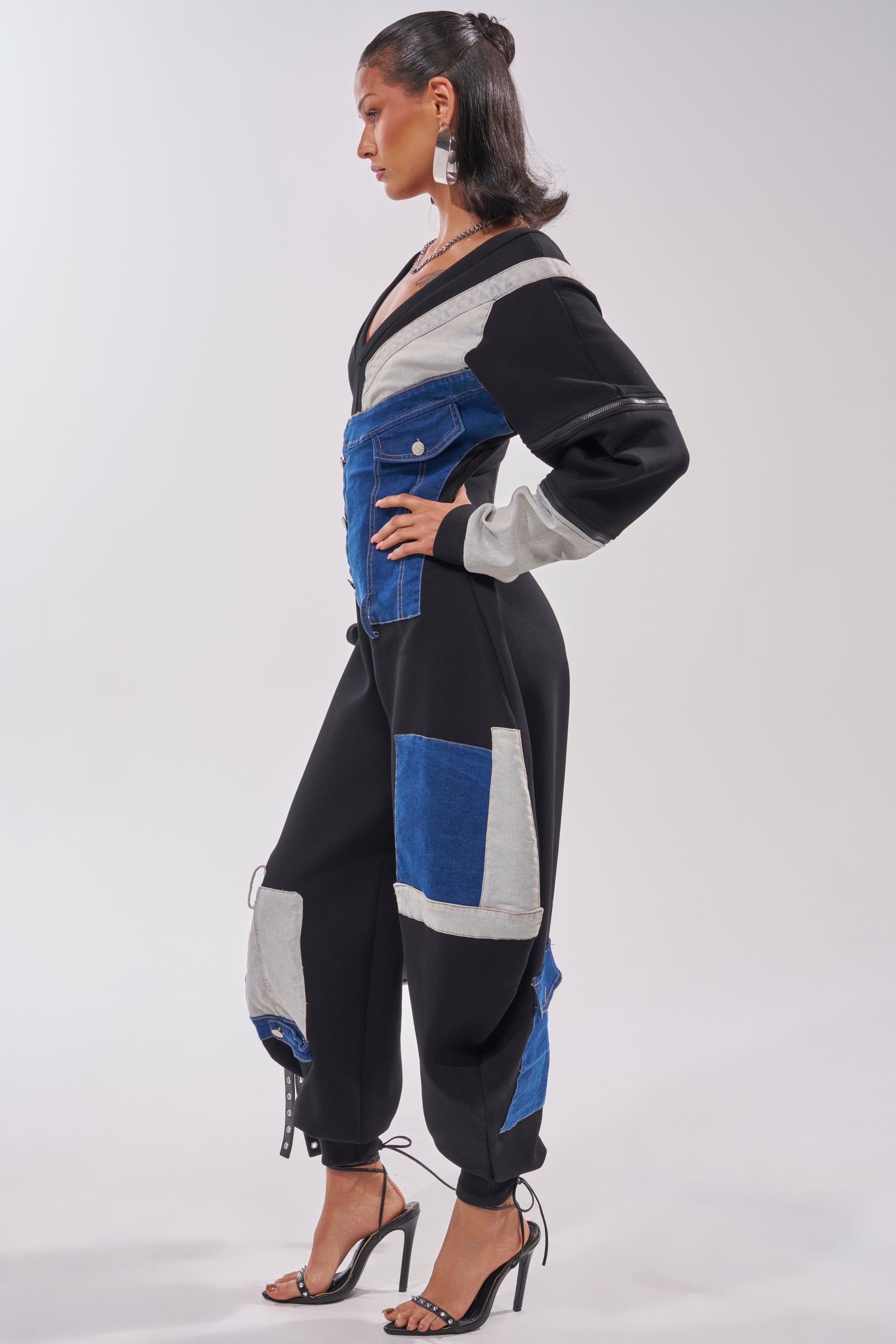 A woman stands in profile wearing the COMFY COZY SWEATSHIRT JUMPSUIT IN PATCHWORK DENIM IN BLACK, featuring black, blue, and gray sections with long sleeves and cargo pockets, paired with black heels for a chic streetwear look. 5