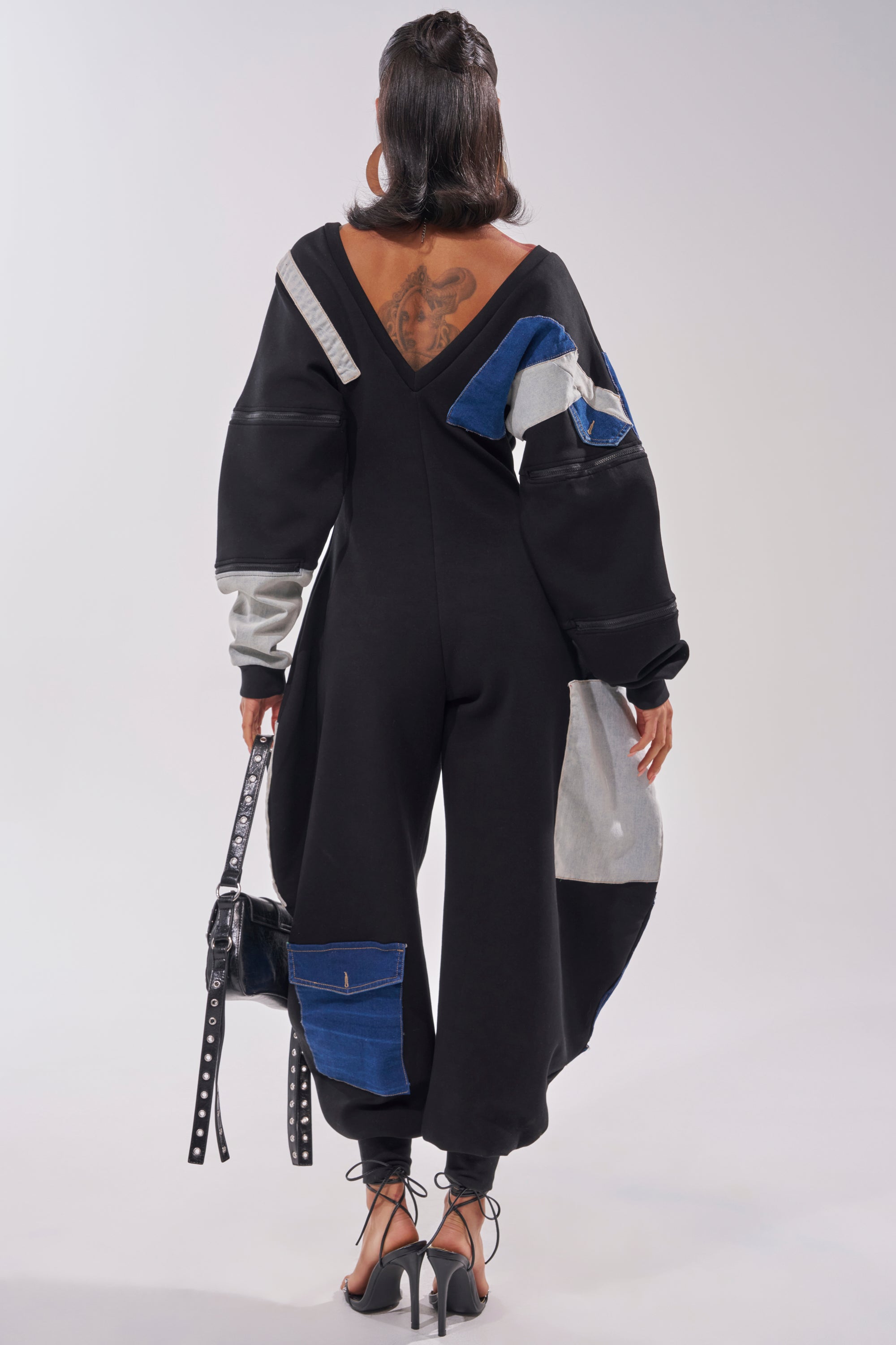 A person with a large back tattoo wears the COMFY COZY SWEATSHIRT JUMPSUIT IN PATCHWORK DENIM IN BLACK, holding a black handbag and standing in strappy heels, facing away from the camera. 6