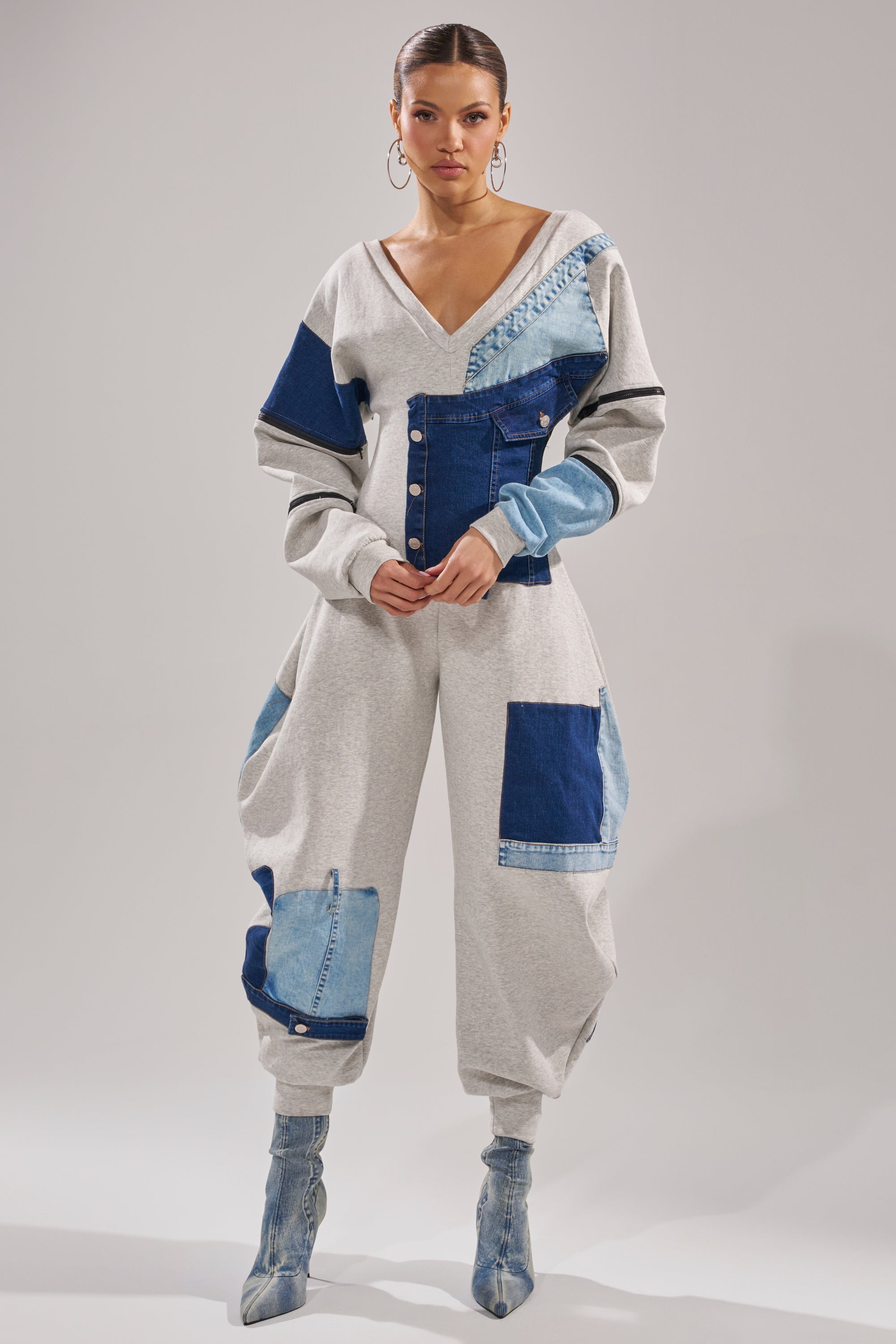 A woman stands confidently in the COMFY COZY SWEATSHIRT JUMPSUIT IN PATCHWORK DENIM, featuring a V-neckline and loose-fitting pants. She styles it with denim ankle boots and large hoop earrings against a plain background.
