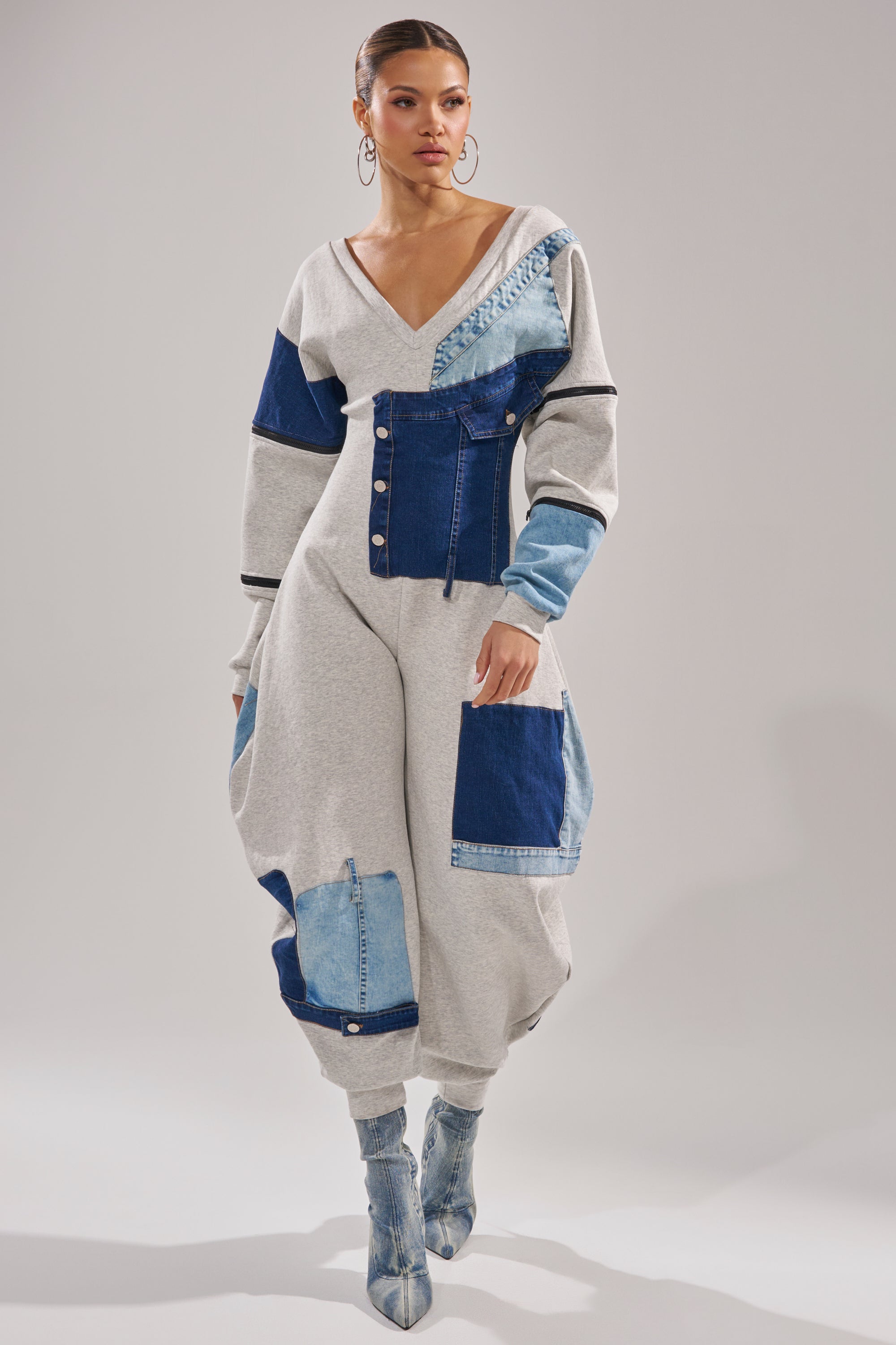 A woman exudes modern streetwear style in the COMFY COZY SWEATSHIRT JUMPSUIT IN PATCHWORK DENIM, with large pockets and blue-heeled boots, bold hoop earrings, and sleek hair against a plain backdrop.