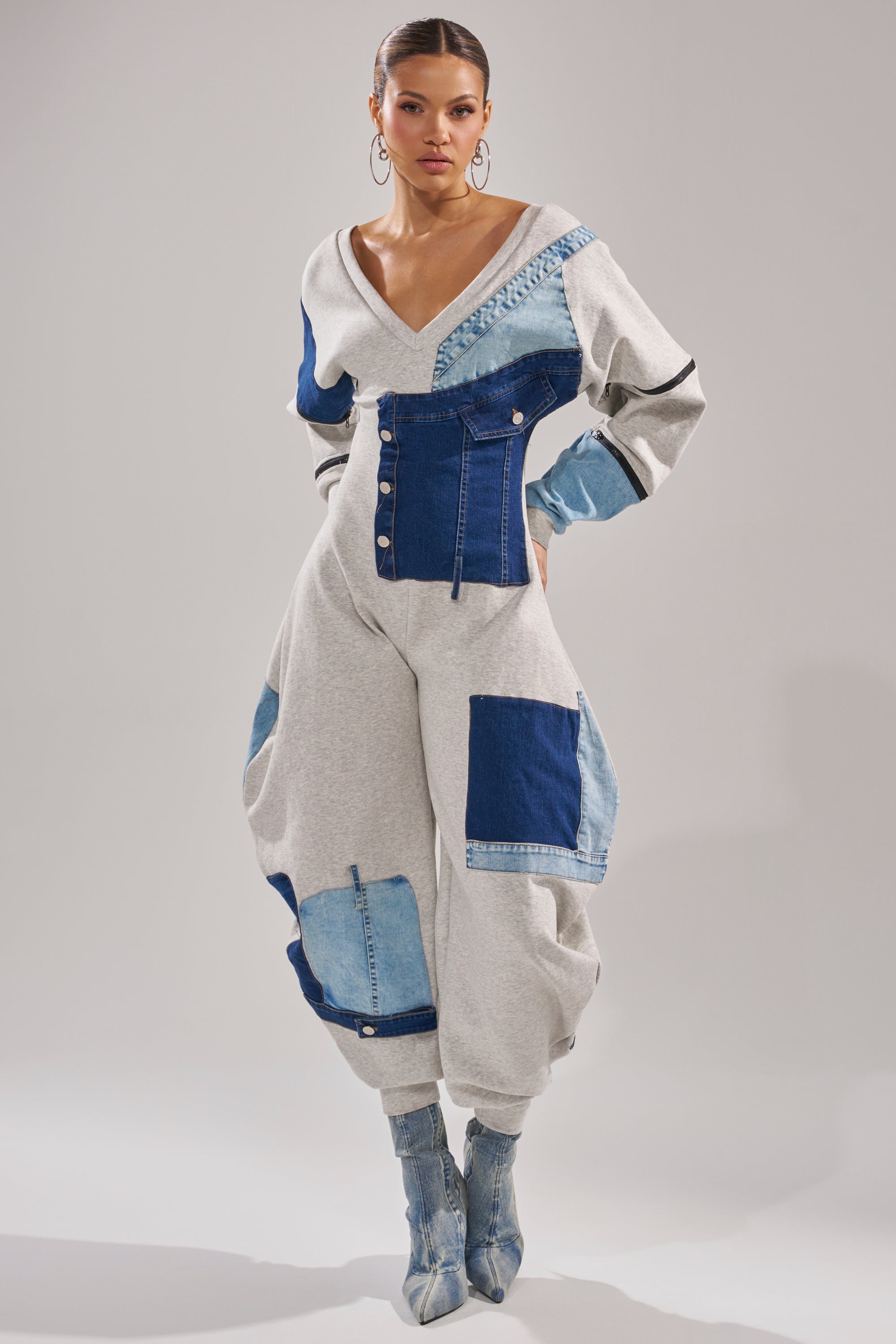 A woman poses in the COMFY COZY SWEATSHIRT JUMPSUIT IN PATCHWORK DENIM, featuring a deep V-neckline, button details, and cuffed ankles. She styles it with denim boots and large hoop earrings against a plain background.