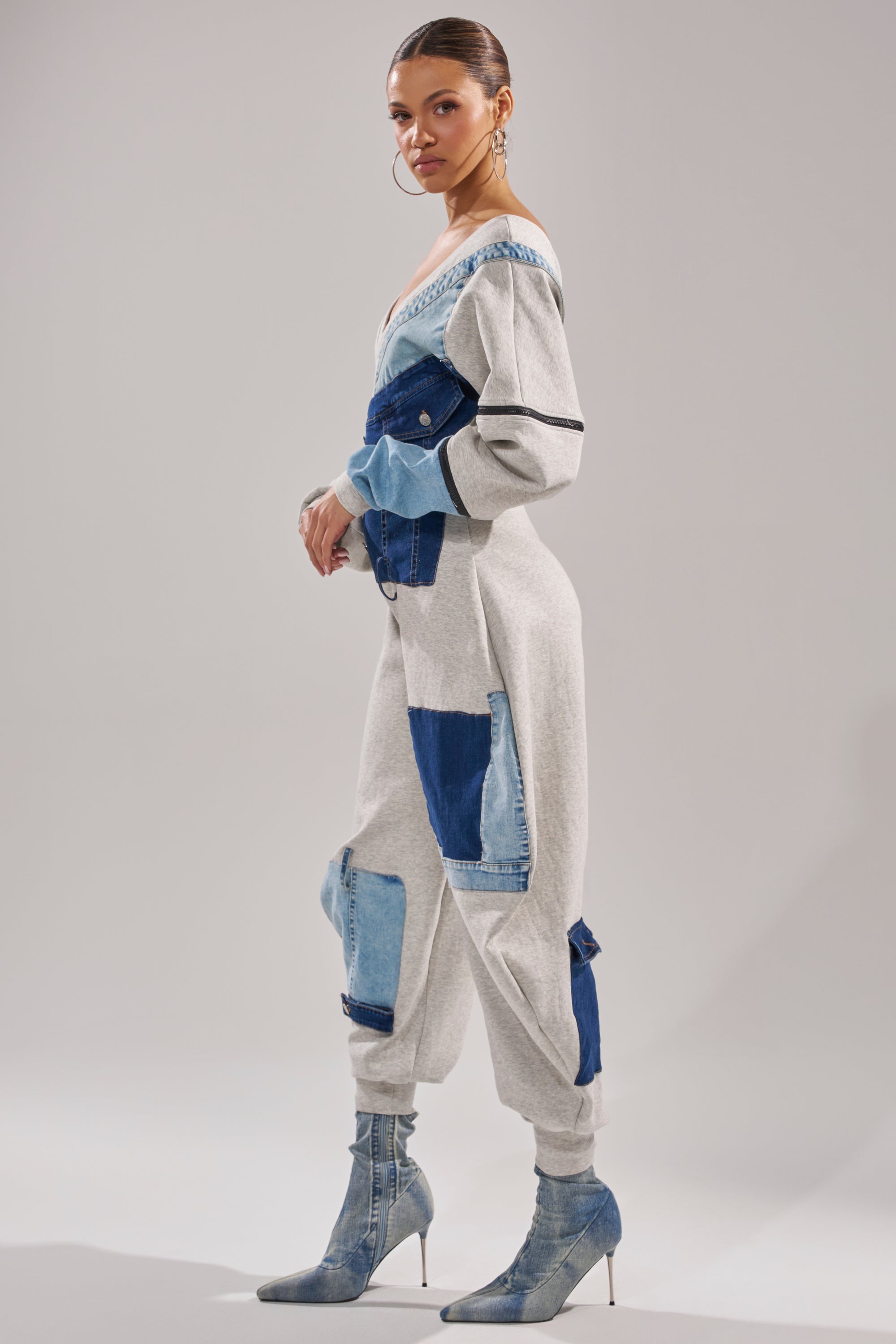 A woman in the COMFY COZY SWEATSHIRT JUMPSUIT IN PATCHWORK DENIM pairs blue heeled ankle boots and large hoop earrings with a sleek bun, showcasing a modern streetwear style against a plain light backdrop.