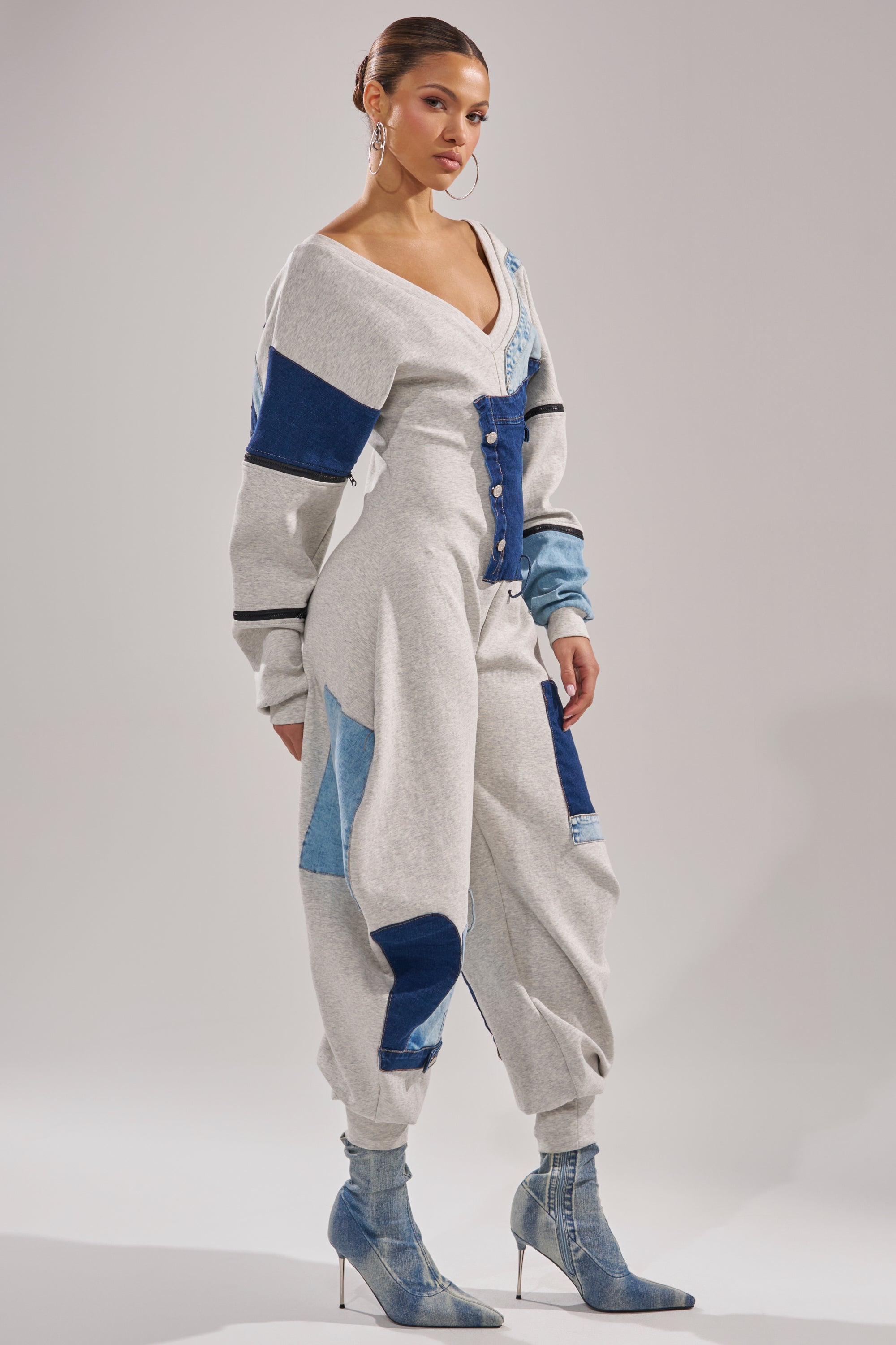 A woman models the COMFY COZY SWEATSHIRT JUMPSUIT IN PATCHWORK DENIM with grey and blue denim patches, button accents, and a deep V-neckline, styled with hoop earrings and pointed toe denim boots against a light background.