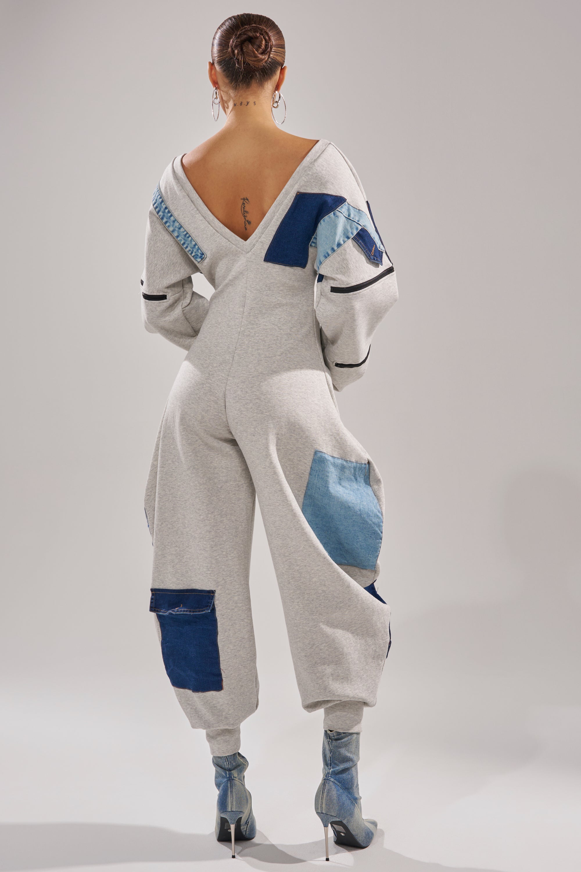 A person faces away in the COMFY COZY SWEATSHIRT JUMPSUIT IN PATCHWORK DENIM, with visible seams, metallic blue high-heeled boots, hair in a bun, and a tattoo showing on their upper back.