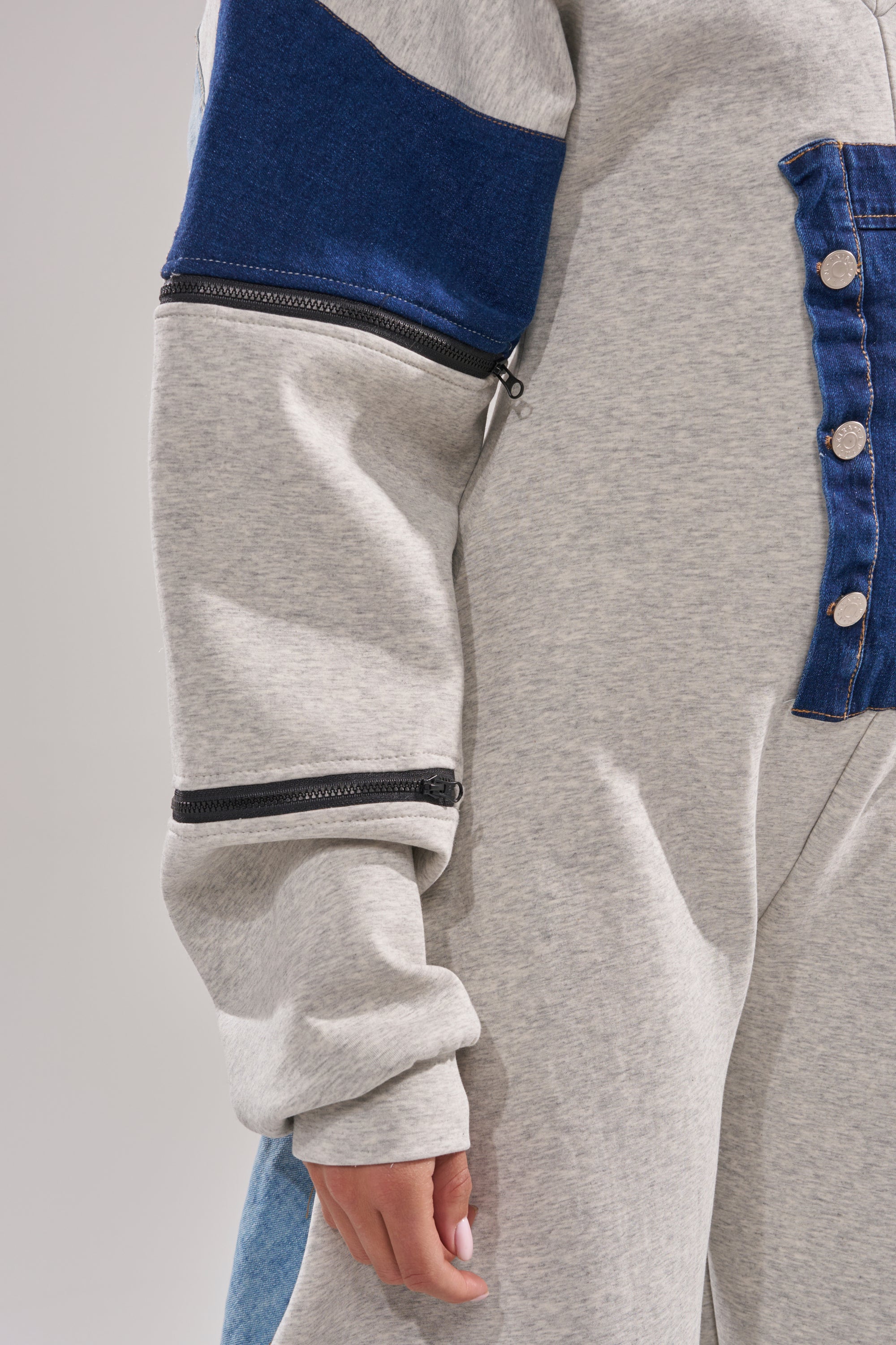 A person wears the COMFY COZY SWEATSHIRT JUMPSUIT IN PATCHWORK DENIM, featuring blue denim patches on the chest and upper arm, metallic buttons, a black sleeve zipper, and a partially visible hand at the bottom.