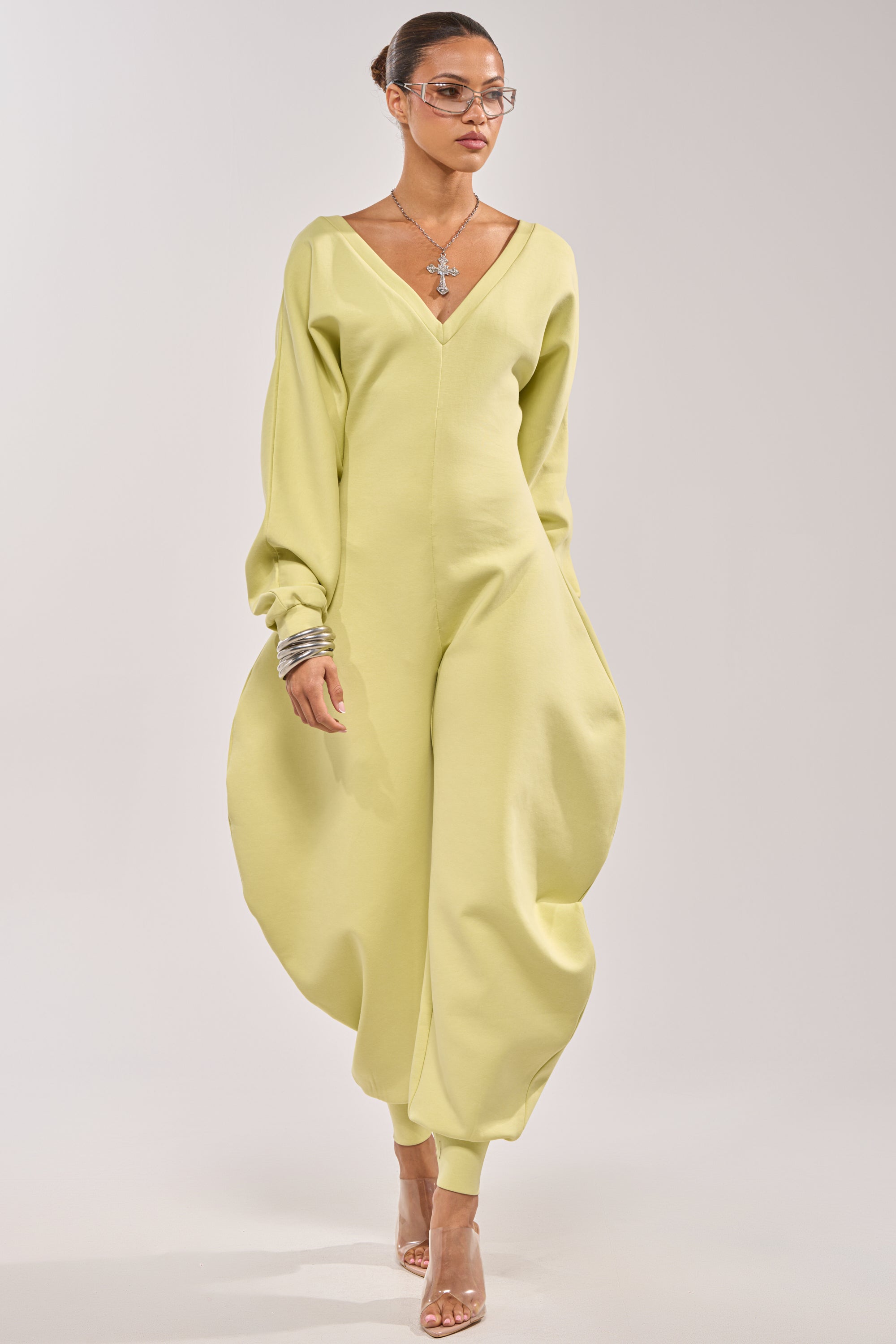 A model showcases the COMFY COZY SWEATSHIRT JUMPSUIT IN LIME, featuring voluminous rounded pant legs, long sleeves, and a deep V-neckline. She styles it with clear heels, layered bracelets, a large necklace, and glasses—a chic lounge look. 0