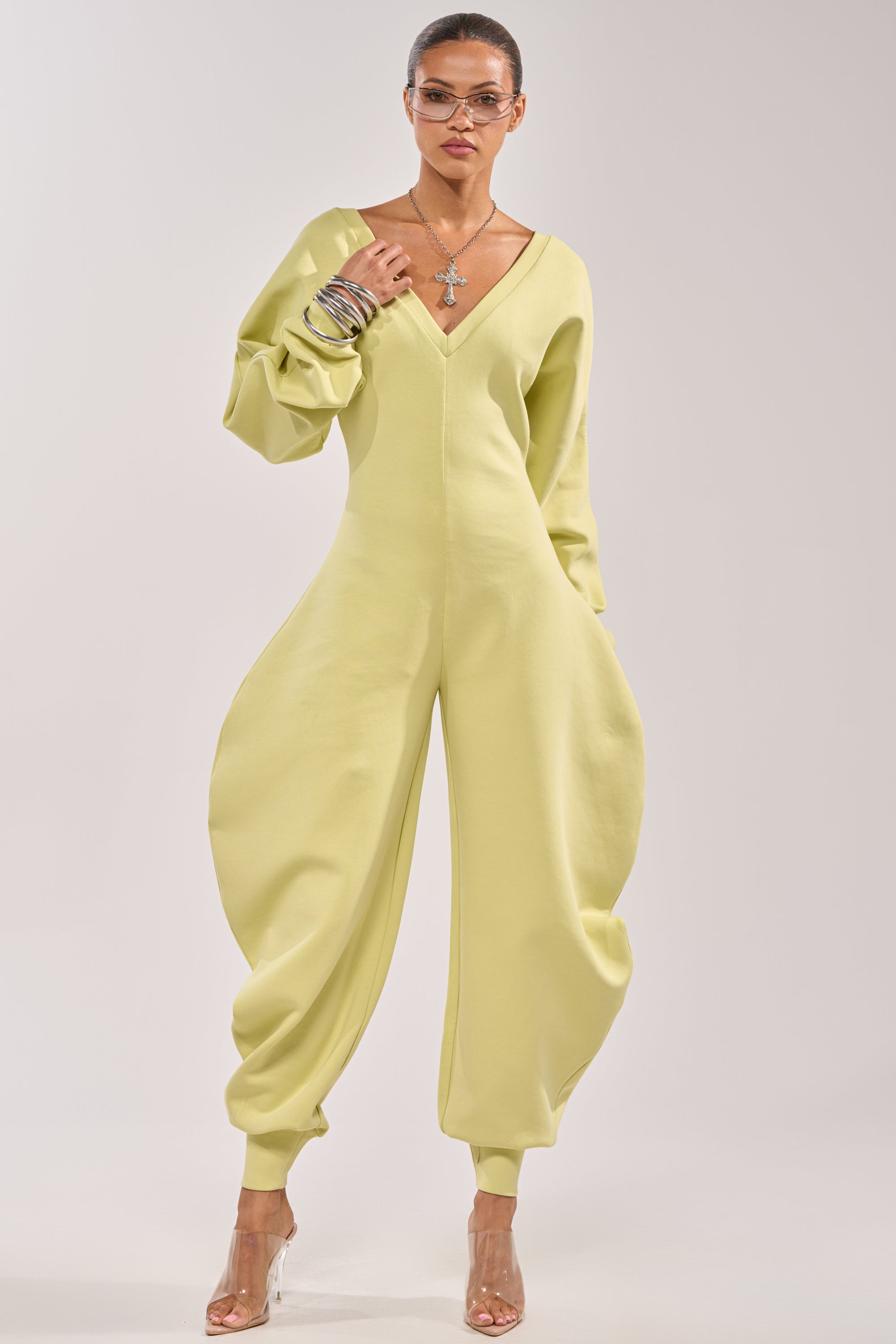 Someone stands confidently against a plain background wearing the COMFY COZY SWEATSHIRT JUMPSUIT IN LIME, featuring a contemporary streetwear look with voluminous legs and sleeves, styled with clear heels and silver jewelry. 1