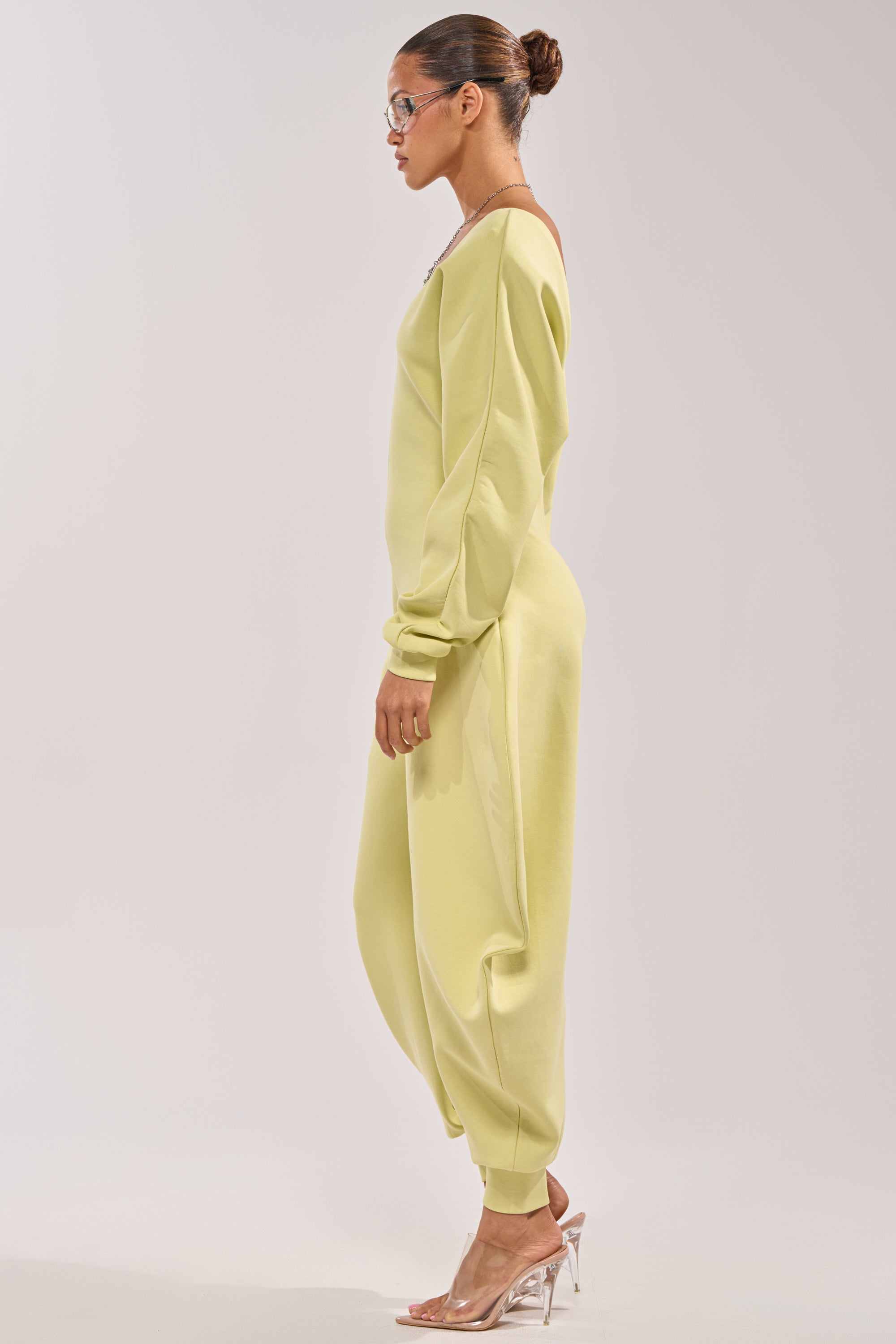 A woman in profile wears the COMFY COZY SWEATSHIRT JUMPSUIT IN LIME with her hair in a bun, glasses, and clear heels, set against a plain light background. 2