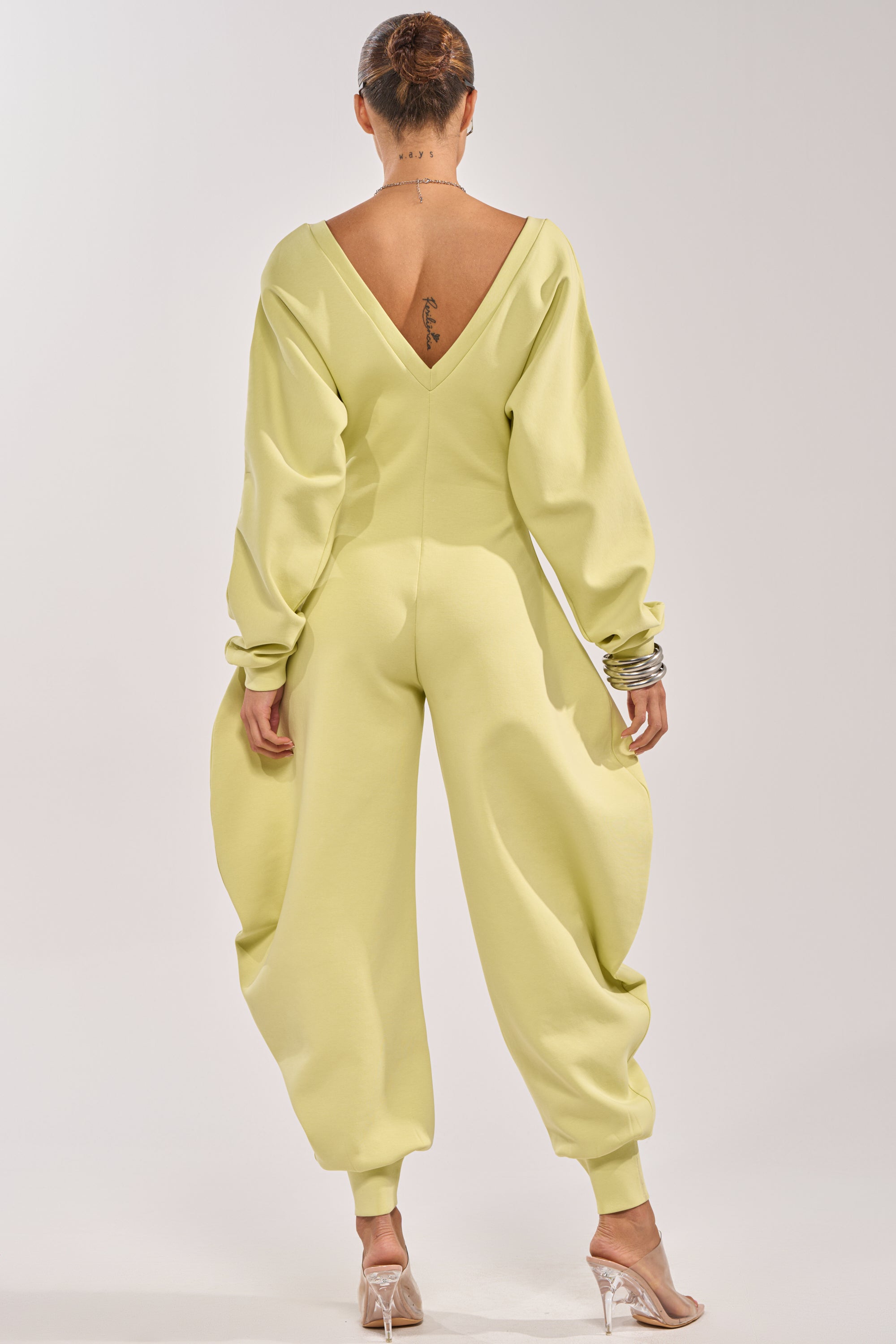 A woman, hair in a bun, wears the COMFY COZY SWEATSHIRT JUMPSUIT IN LIME featuring a deep V-back and voluminous sleeves and legs, styled with clear high heels for a modern streetwear look. 3