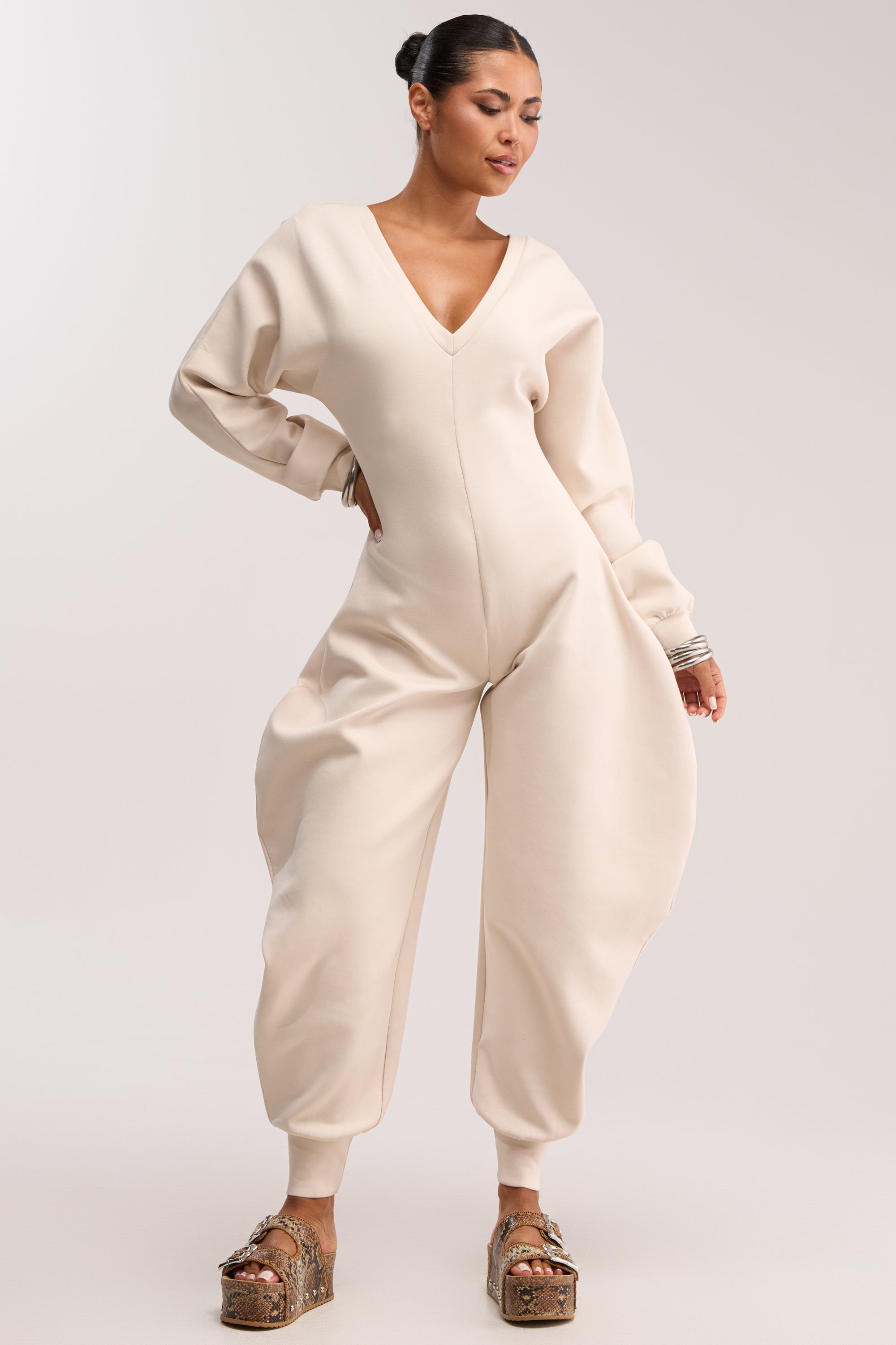 A woman stands confidently in the COMFY COZY SWEATSHIRT JUMPSUIT IN IVORY, featuring exaggerated rounded hips and a V-neckline. Her hair is in a bun, styled with platform sandals and bracelets, set against a plain light background.