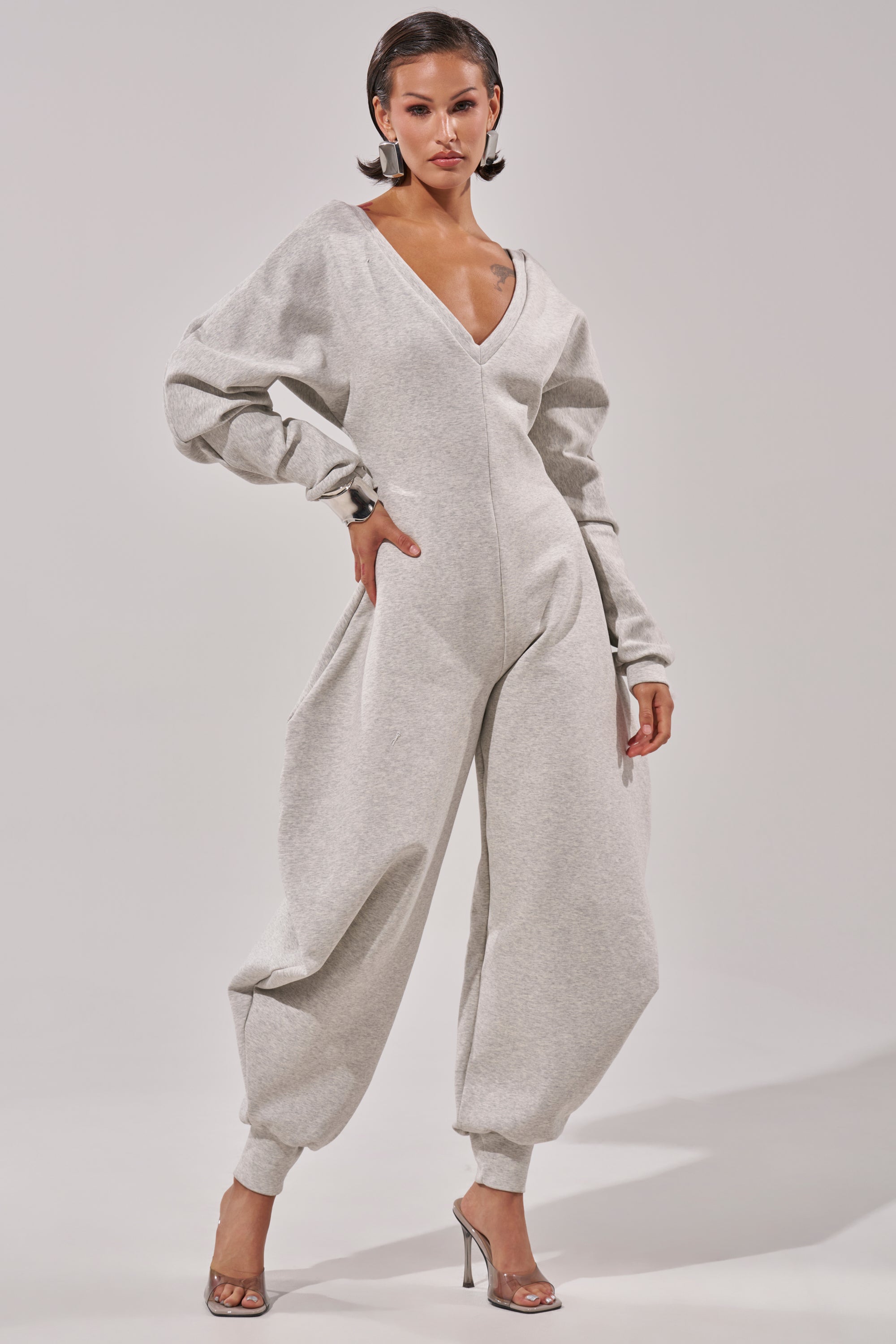 A woman poses confidently in the COMFY COZY SWEATSHIRT JUMPSUIT, a light gray, long-sleeve cotton jumpsuit with a deep V-neck, styled with clear high-heeled sandals against a plain white background.