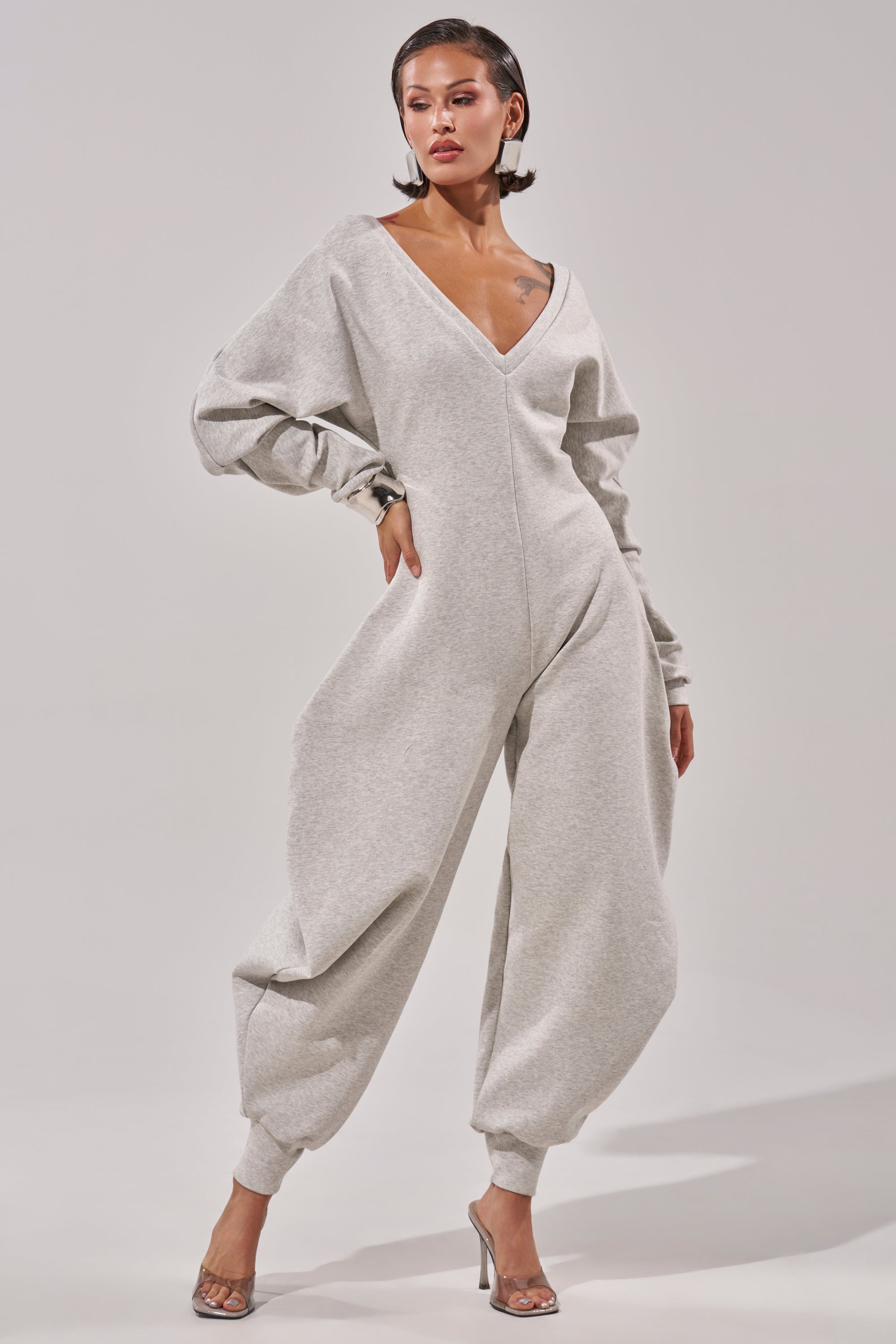 A woman models the COMFY COZY SWEATSHIRT JUMPSUIT, a light gray oversized outfit with long sleeves and baggy legs, styled with clear high heels against a plain light background.