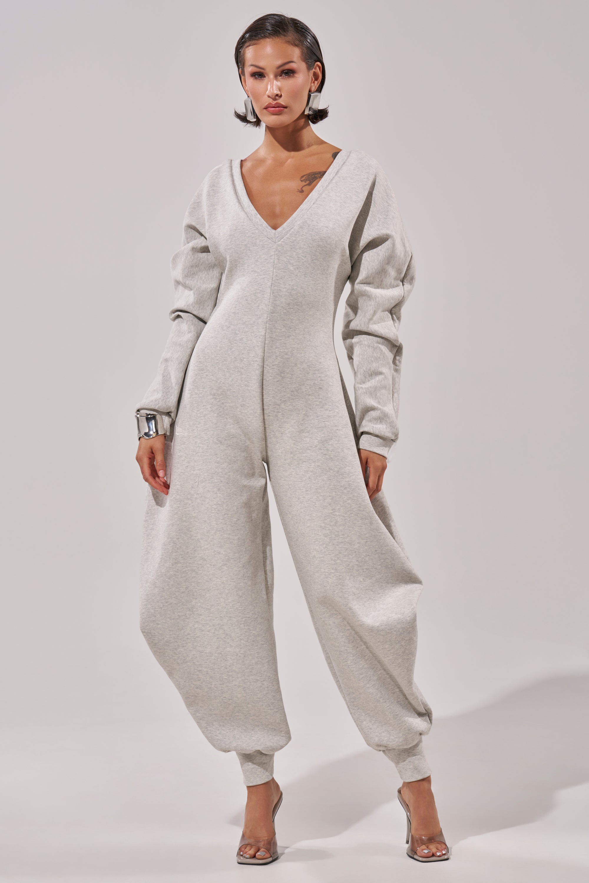 A woman models the COMFY COZY SWEATSHIRT JUMPSUIT in light gray, featuring long sleeves and loose ankle-gathered legs. She pairs it with clear heels and a chunky bracelet, standing against a plain background for a relaxed look.