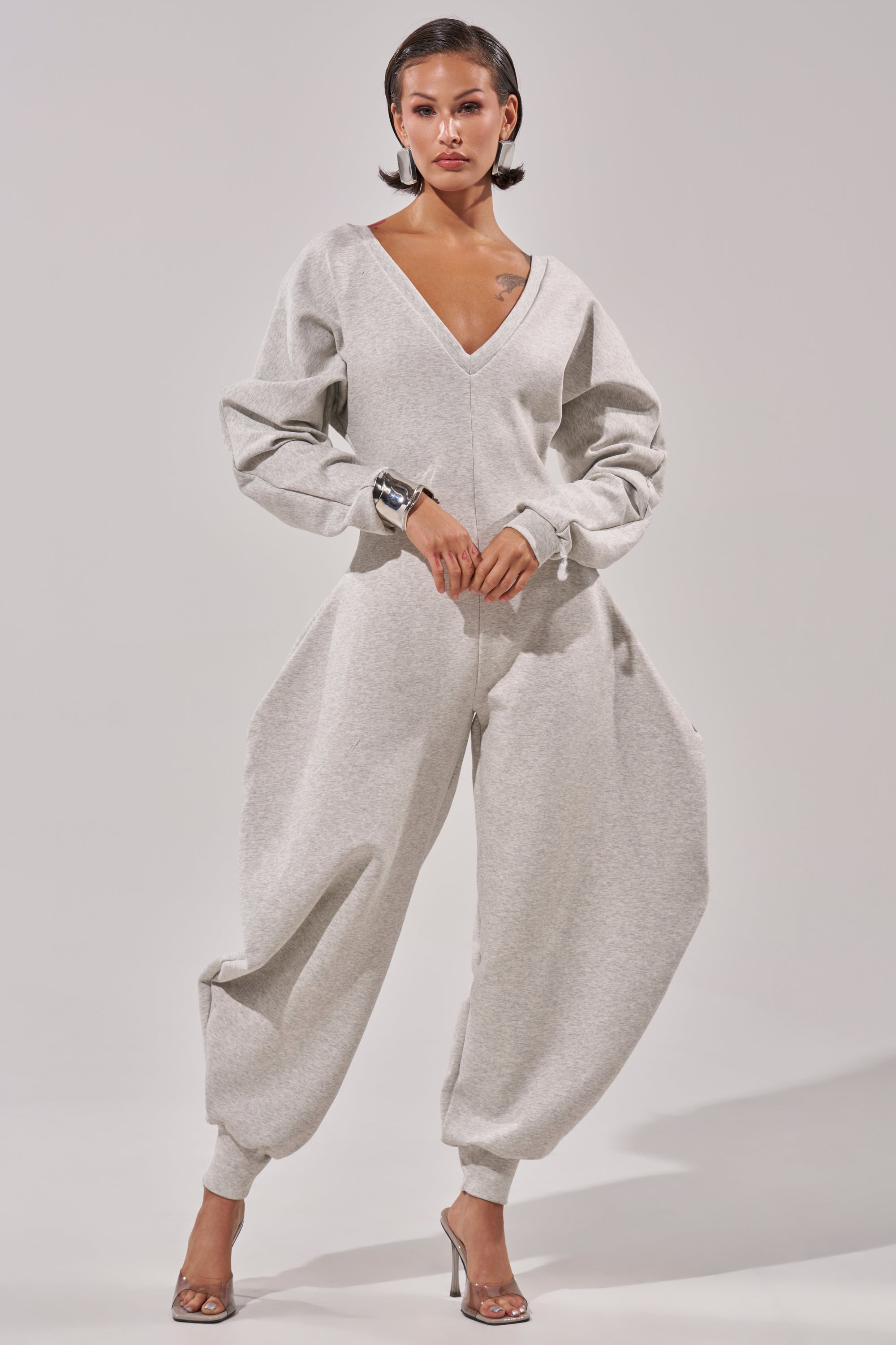 A woman poses in the COMFY COZY SWEATSHIRT JUMPSUIT, a light grey cotton piece with balloon sleeves and wide legs. She pairs it with silver high-heeled sandals and a chunky bracelet, standing against a plain light background.