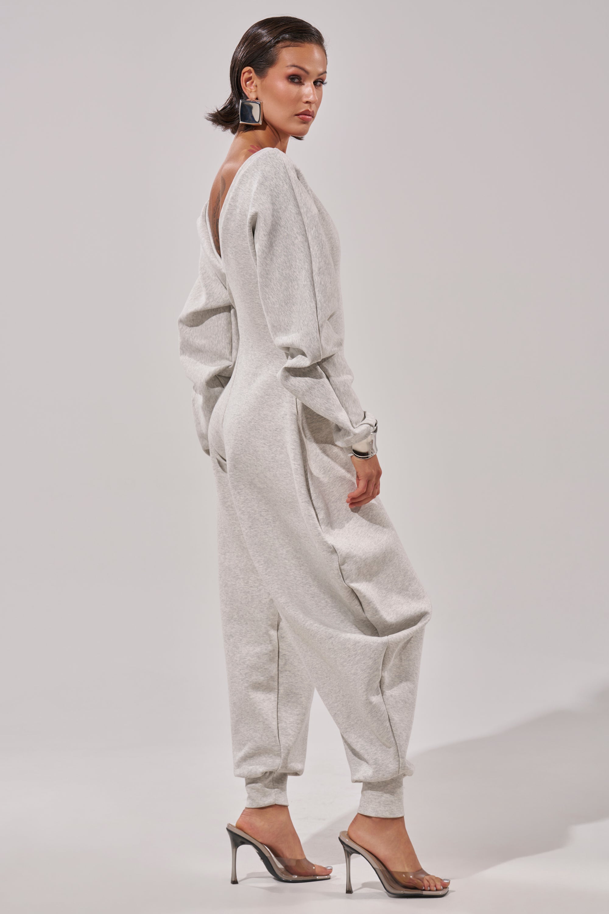 A woman models the COMFY COZY SWEATSHIRT JUMPSUIT in light gray, featuring long sleeves and a backless design, paired with clear high-heeled sandals, standing in a studio with a neutral expression for a relaxed vibe.