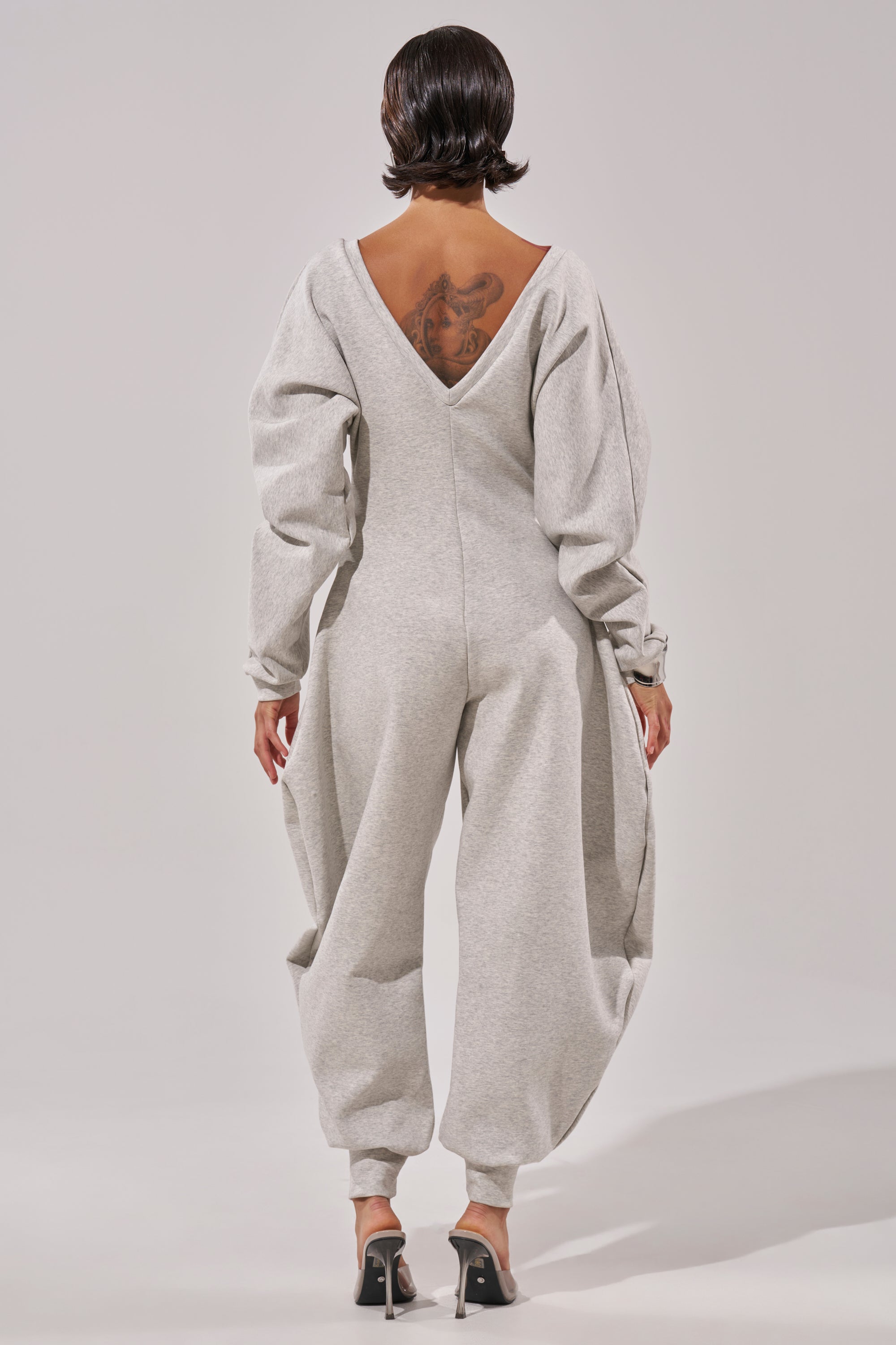 A person with short, dark hair, tattoo on upper back, stands facing away in the COMFY COZY SWEATSHIRT JUMPSUIT—a light gray cotton piece with balloon sleeves and deep V-back—paired with clear-heeled shoes.