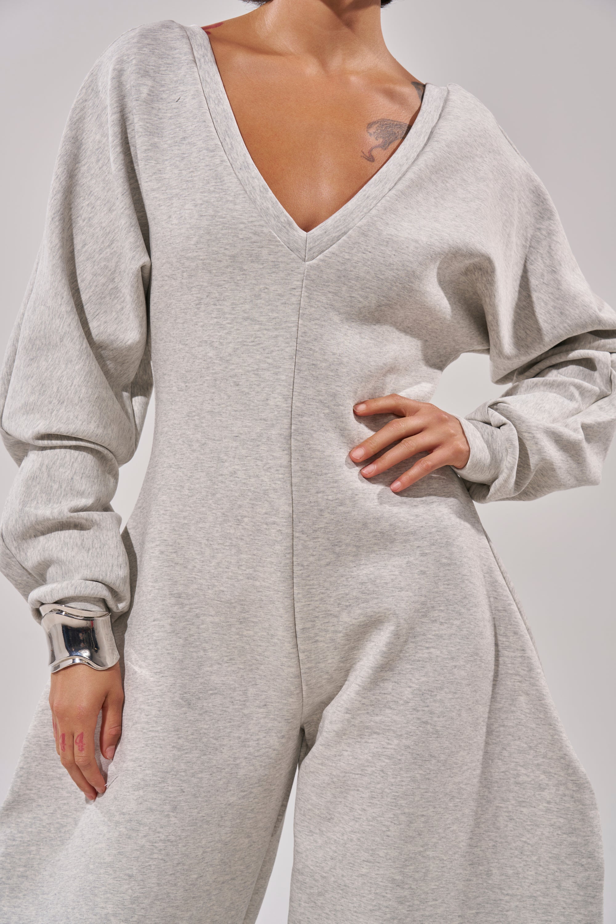 A person models the COMFY COZY SWEATSHIRT JUMPSUIT in light gray, featuring long sleeves and a V-neck. The outfit looks comfortable, with only the lower face and upper body visible; one hand rests on their hip.