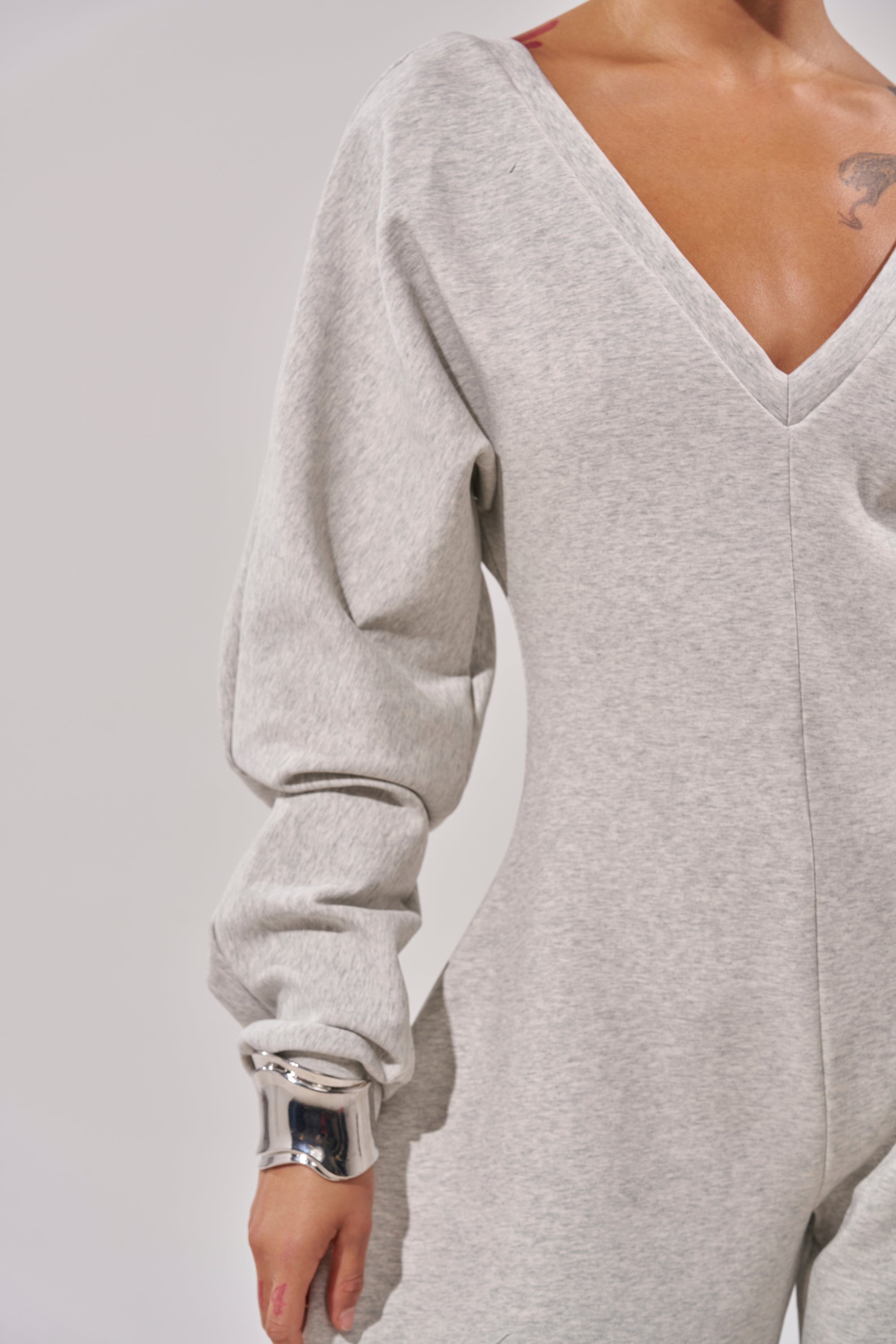 A person models the COMFY COZY SWEATSHIRT JUMPSUIT in light gray with long puffy sleeves, silver bangles on their wrist. The focus is on the upper body, set against a plain, light background for a relaxed look.