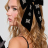 A woman with long curly hair wears the BIG BOW WITH RHINESTONES IN BLACK, an oversized velvet hair clip, adorning the back of her head while she looks to the side against a plain background.