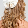 A person with wavy, blonde hair styled with the WINTER WHITE BLING HAIR BOW, featuring satin fabrication and adorned with pearl bead embellishments and cascading chains at the back, against a plain and neutral background.