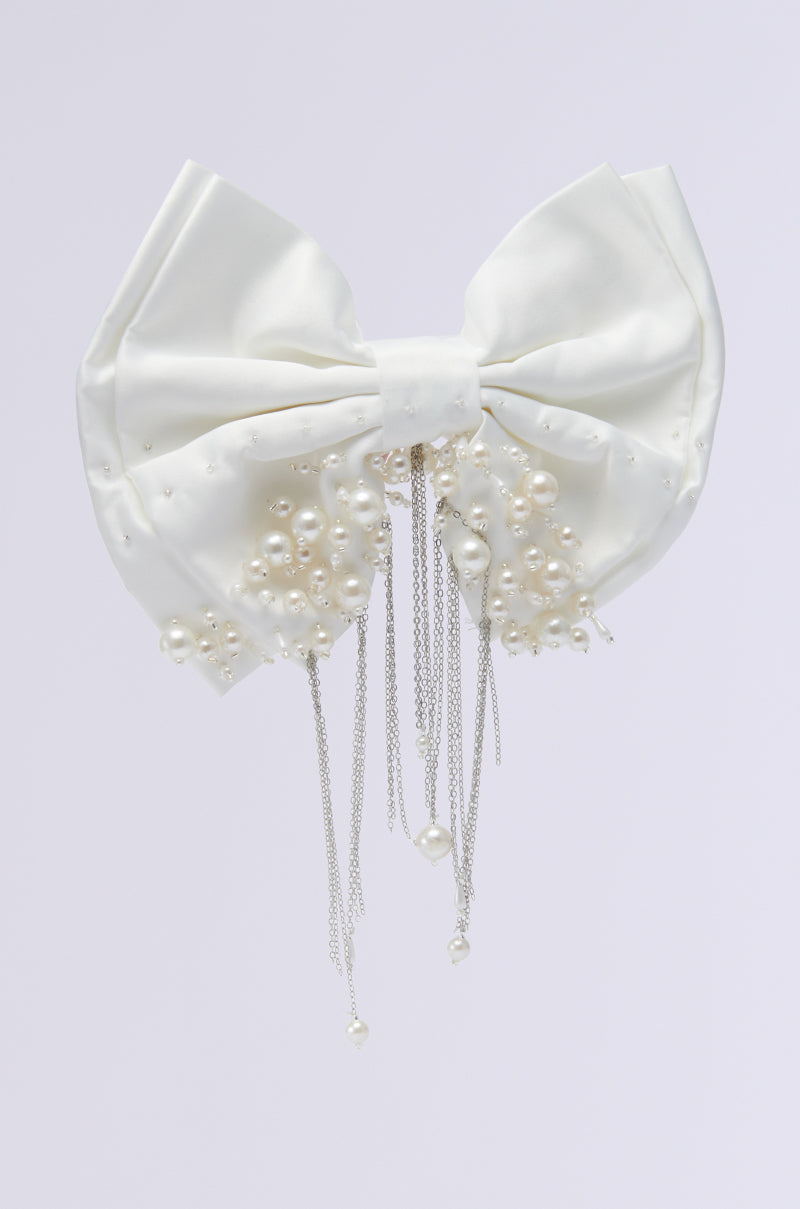 The WINTER WHITE BLING HAIR BOW is an exquisite hair accessory made from luxurious satin, featuring a large white satin bow decorated with dangling silver chains and pearl bead embellishments on a light gray background. 1