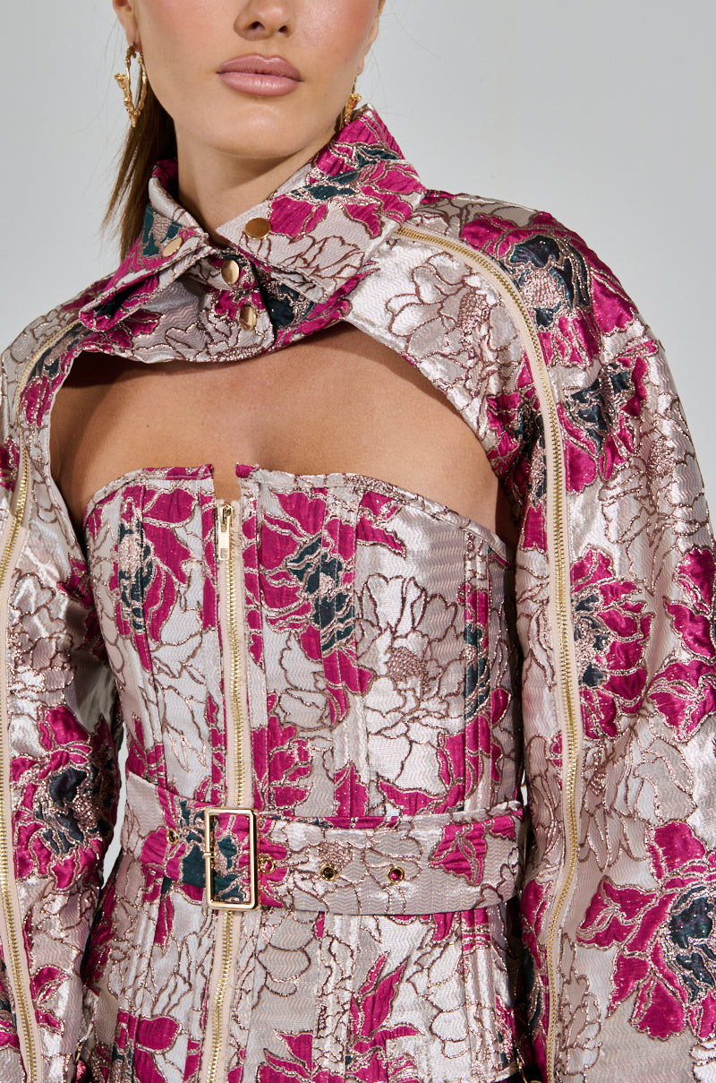 A woman wears the elegant AURORA BROCADE BOLERO, an off-shoulder floral piece in pink, silver, and black with a textured pattern and metallic detailing. The fitted waist zip detail enhances the look, complemented by gold earrings against a light-colored backdrop.