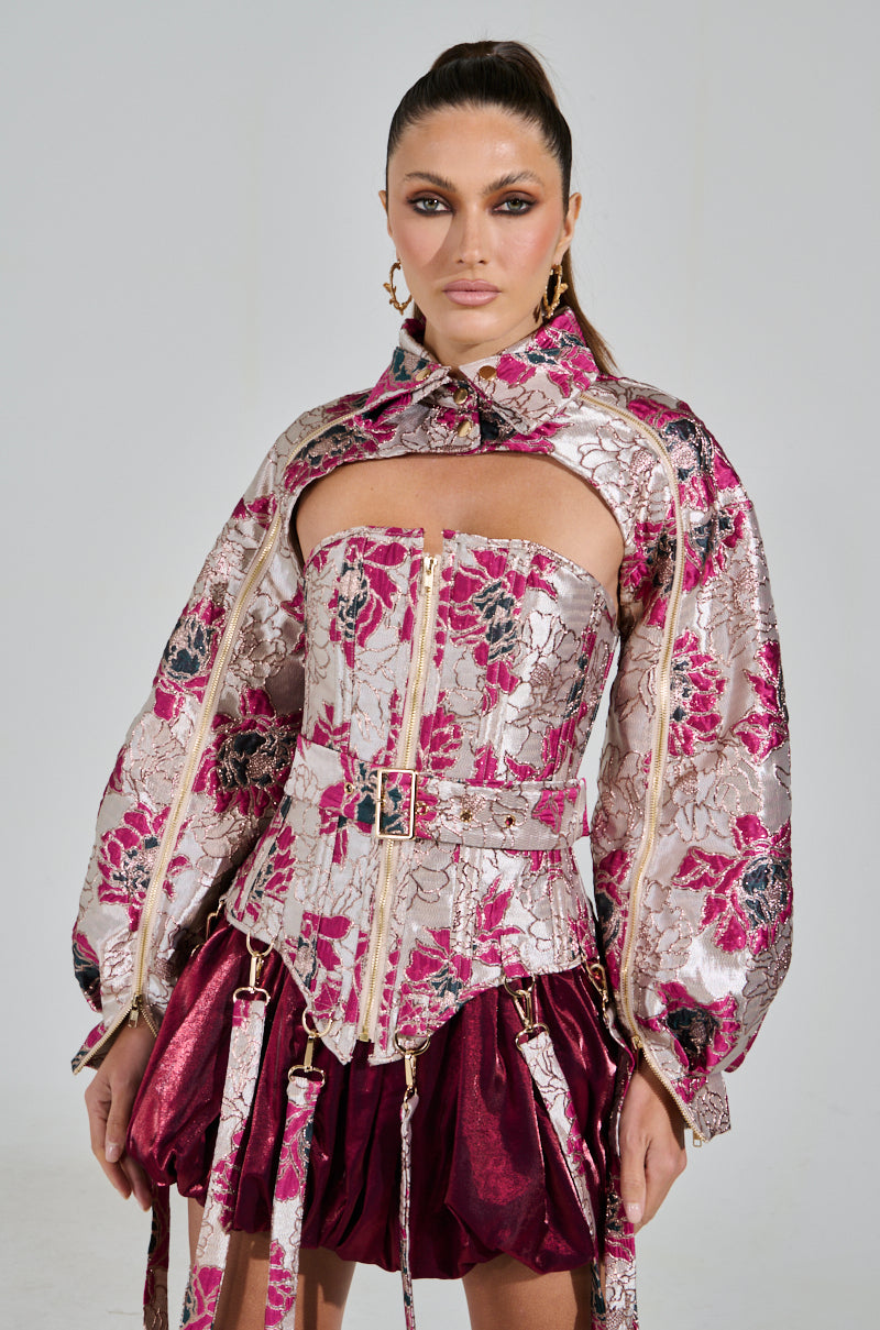 A person wears an AURORA BROCADE BOLERO with ornate floral brocade patterns and metallic details, featuring long sleeves and a high collar. Complemented by a structured corset and skirt in burgundy and silver, it stands elegantly against a plain backdrop.