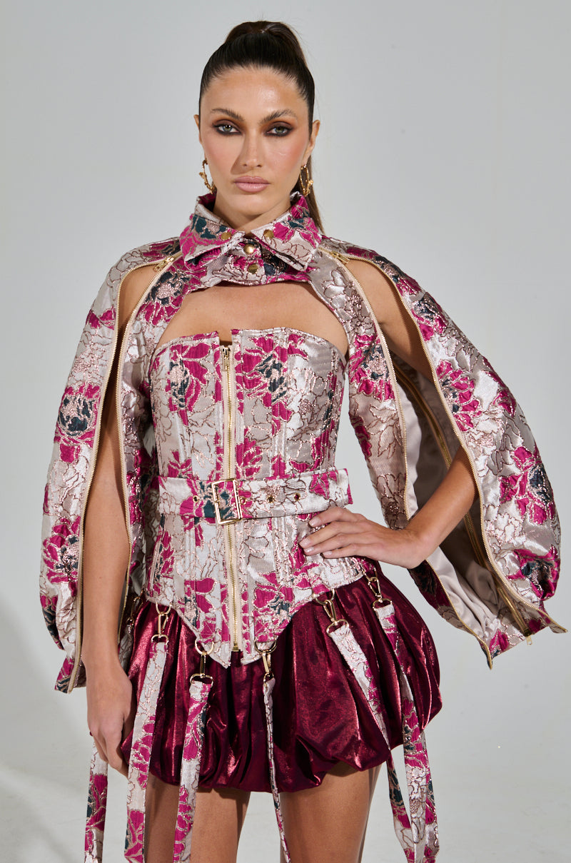 The model showcases the chic AURORA BROCADE BOLERO, matched with a floral pattern corset and pleated skirt adorned with dark red accents. Her hair is slicked back, bold makeup perfects the look against a stark background.