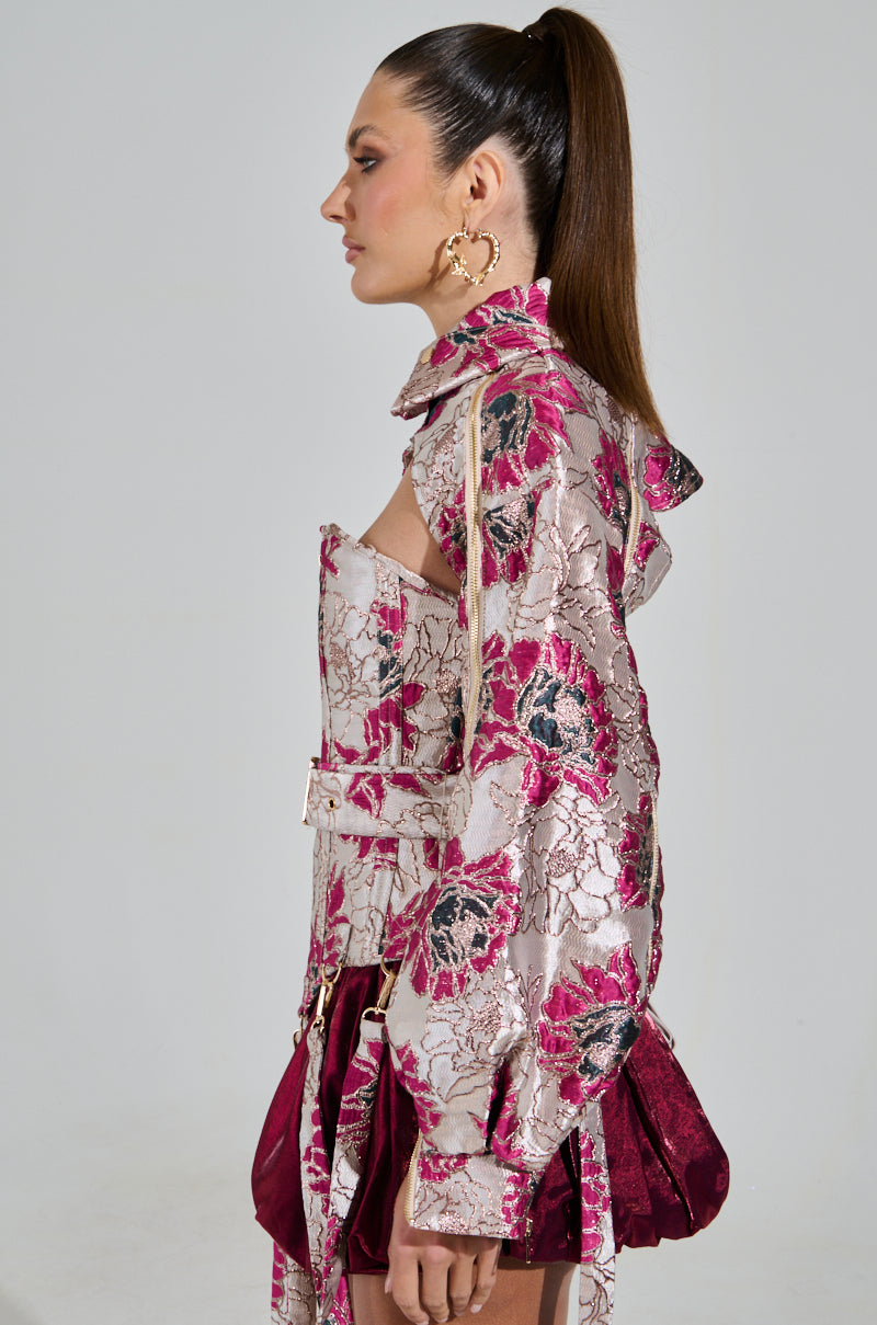 A woman with a high ponytail wears the chic AURORA BROCADE BOLERO, an off-shoulder top in pink and silver floral brocade with metallic detailing and ruffle accents. She complements her look with large hoop earrings against a neutral backdrop.