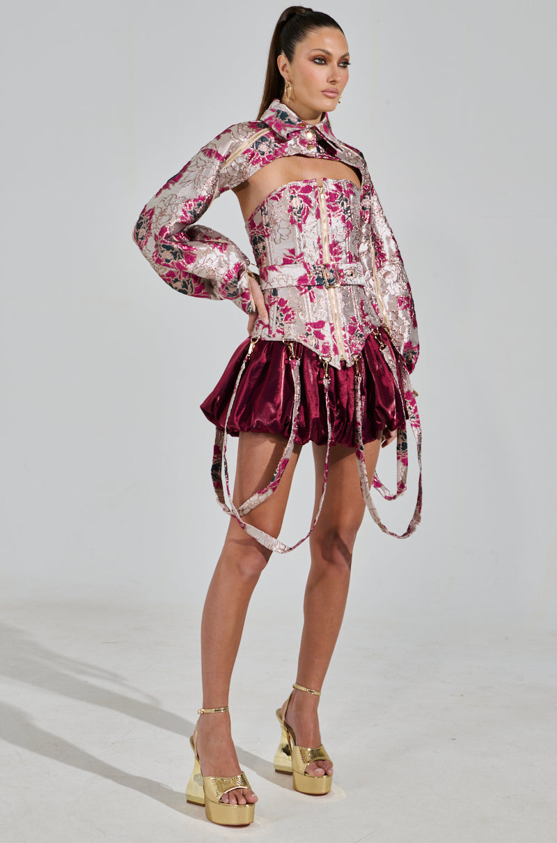 A model showcases a fashion-forward look in the Aurora Brocade Bolero, a floral brocade-patterned top with an exposed midriff and long sleeves, paired with a burgundy velvet skirt featuring flowing fabric strips, gold platform heels, and metallic detailing for added glam.