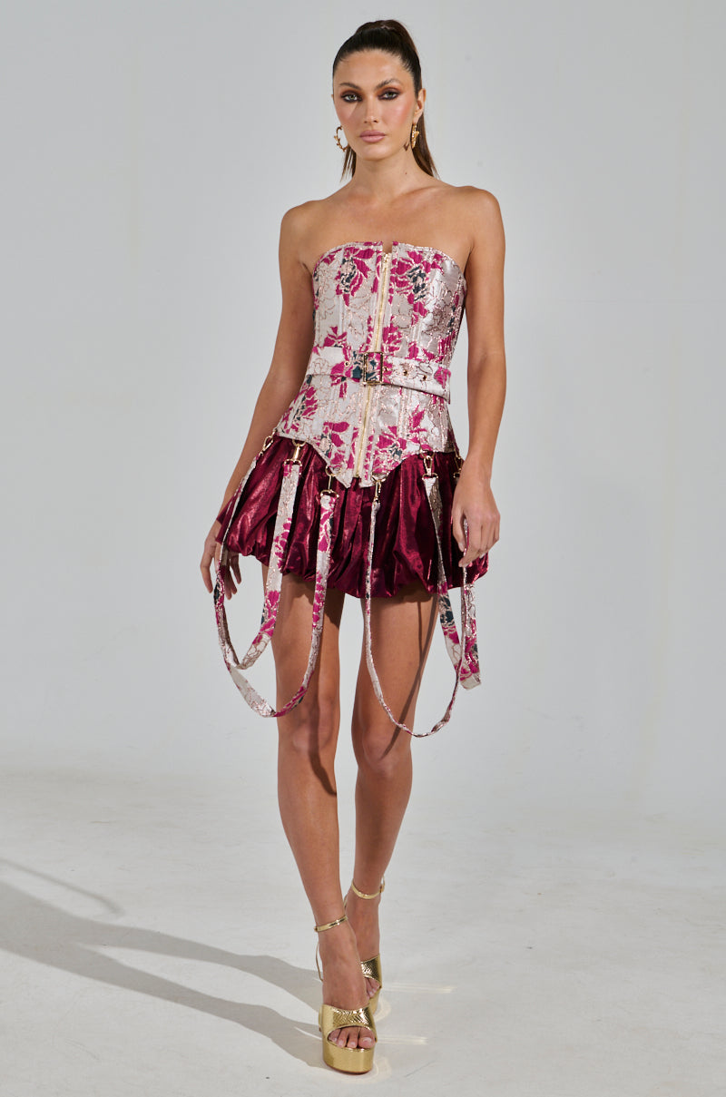 A woman in the Aurora Brocade Corset featuring a floral brocade pattern is elegantly poised in gold high heels against a plain background, paired with a shiny, burgundy pleated skirt with metallic detailing.