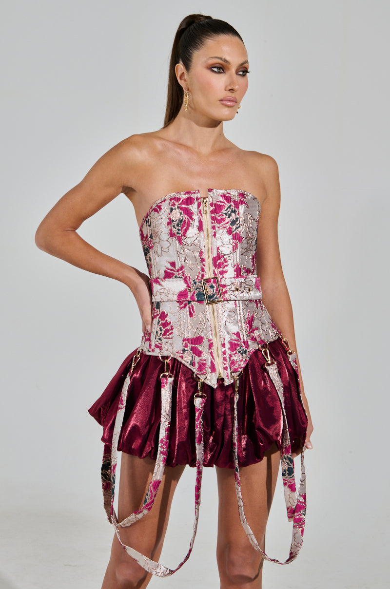 A woman exudes elegance and poise in an AURORA BROCADE CORSET with a floral design, paired with a shiny burgundy pleated skirt. Her hair is styled in a high ponytail as she gazes to the side, one hand resting on her hip.