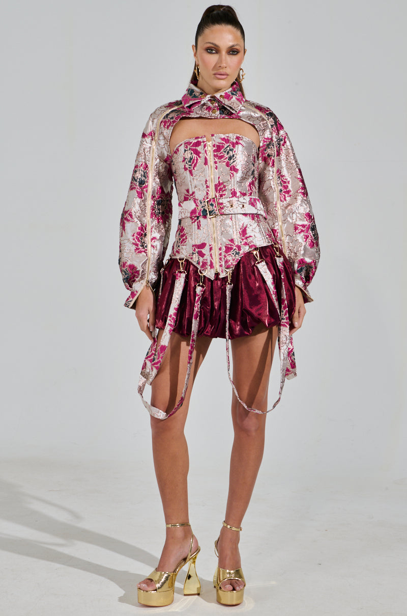 A model wears a floral metallic top with voluminous sleeves and cutouts, similar to an AURORA BROCADE CORSET, paired with a skirt that has metallic suspenders. She completes the look with gold platform shoes and a sleek ponytail against a plain white background.