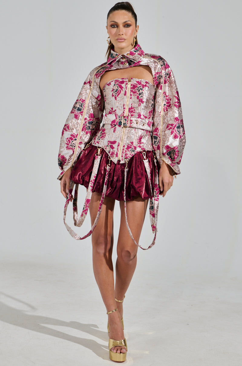 A model showcases the AURORA BROCADE CORSET with dramatic sleeves, paired with a burgundy mini skirt. She completes the look with gold high-heeled sandals featuring metallic details and a choker, all set against a plain backdrop.