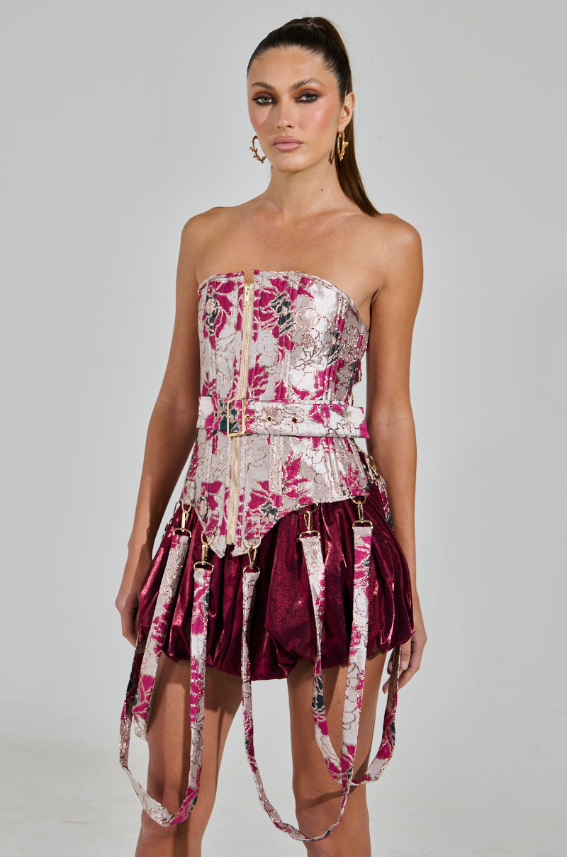 A model poses in a strapless burgundy dress with a structured floral brocade bodice. The Aurora Brocade Corset has multiple buckle straps and metallic details. She accessorizes with gold hoop earrings, long hair, and a confident expression.