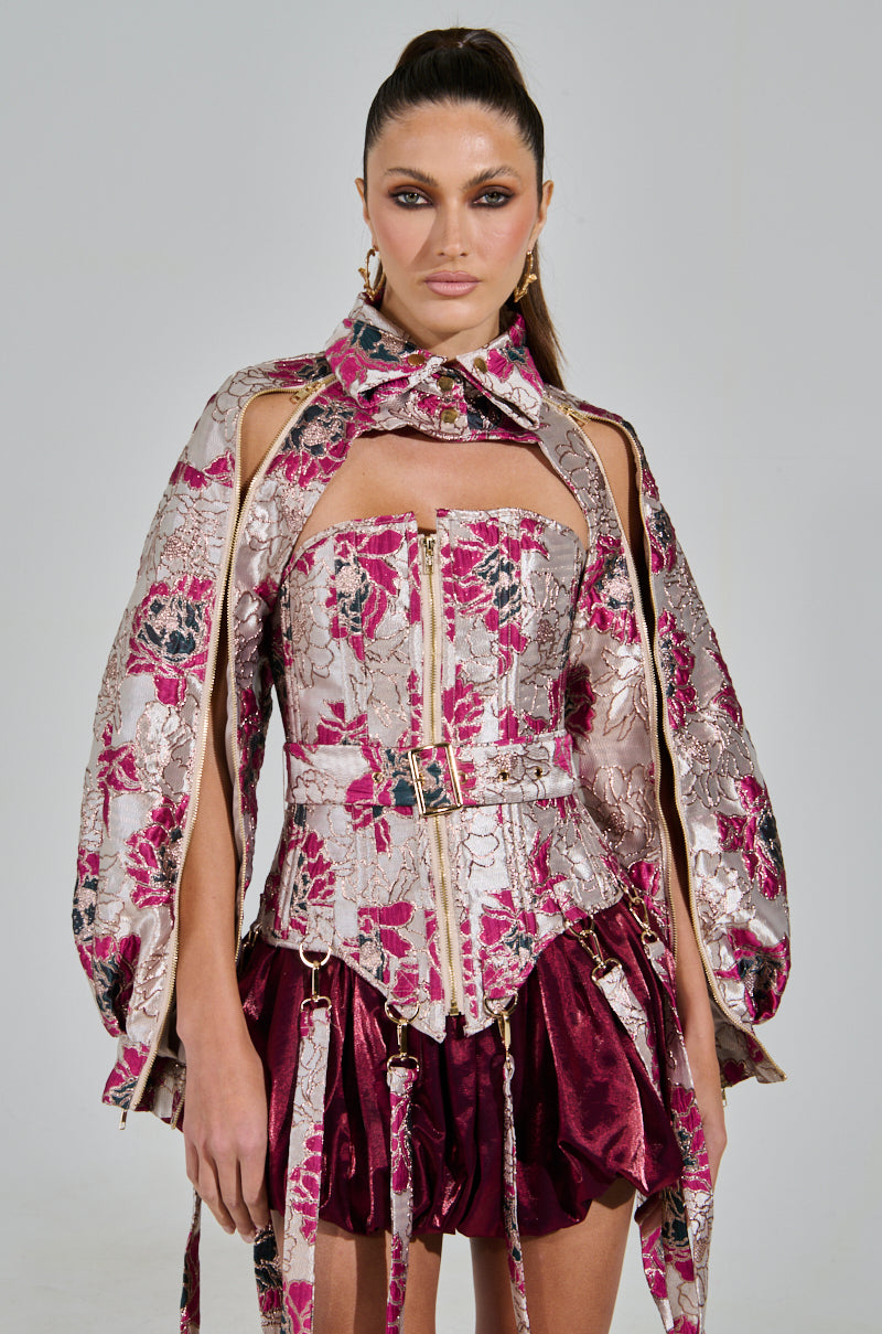 A woman models a chic outfit with the AURORA BROCADE CORSET featuring metallic detailing and a floral brocade pattern. It includes structured shoulders, intricate cutouts, and a matching belt, paired with a short burgundy skirt. Her hair is styled back, complemented by bold makeup.