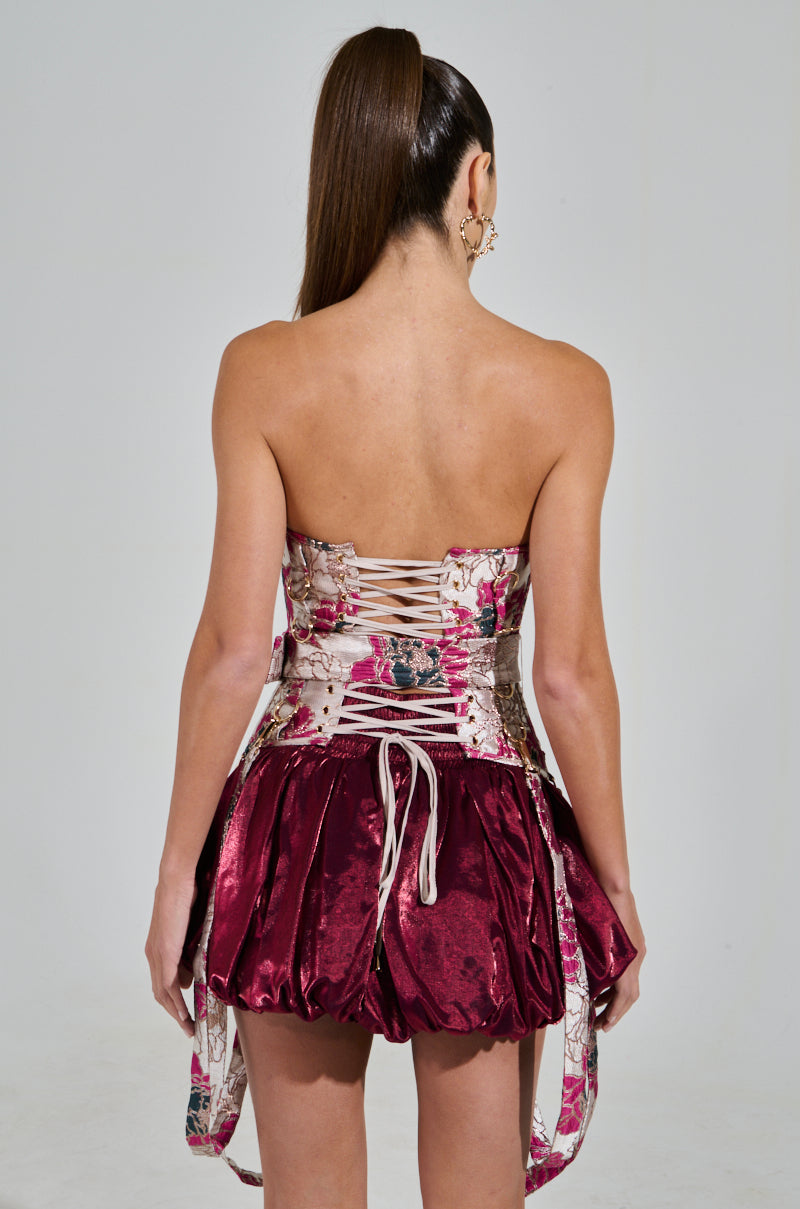 A woman with long hair in a high ponytail, facing away, wears an AURORA BROCADE CORSET dress. The strapless design features a floral brocade pattern, lace-up detailing, and a short, voluminous satin skirt. She stands against a plain gray background.