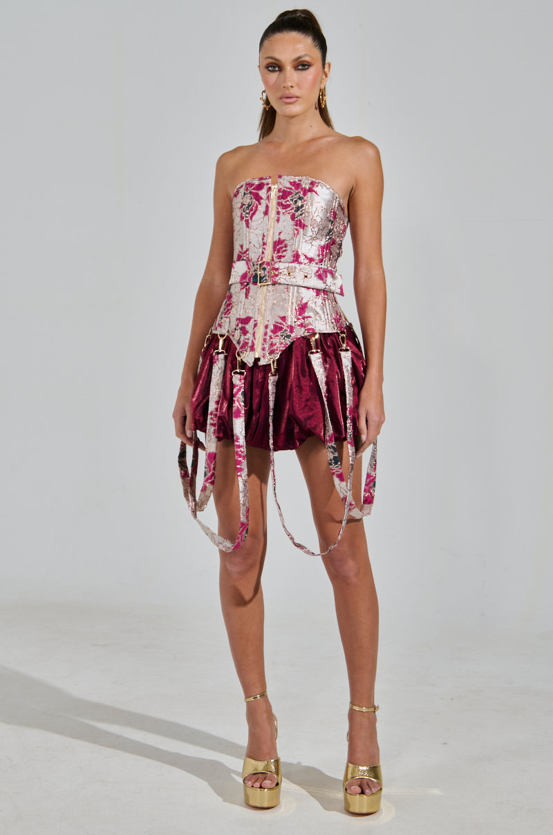 A confident model stands against a plain background, styled with neat hair and wearing the strapless AURORA BROCADE CORSET featuring a floral pattern and metallic detailing. She pairs it with a maroon skirt and gold platform heels.