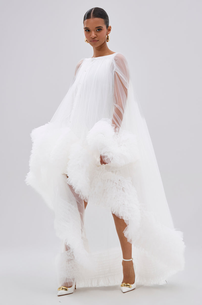 A woman poses in the PIPER SOFT TULLE BLOUSE IN WHITE, a sheer top with long transparent sleeves and ruffled trim, styled with gold earrings and white heels against a plain light background.